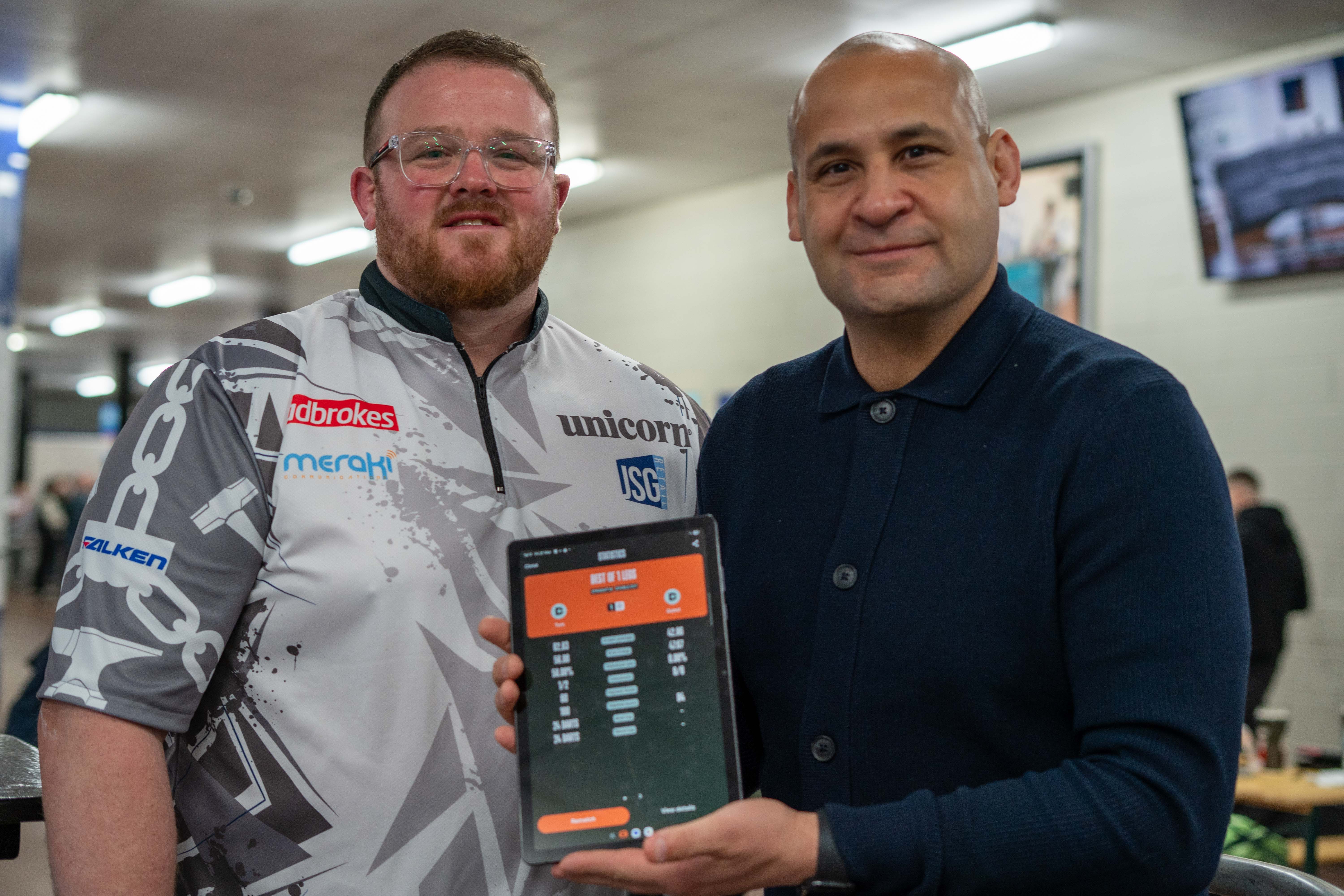 PDC Pro Dart player Tom Bissell & Nigel Quashie holding up a tablet with the score of their match displayed.