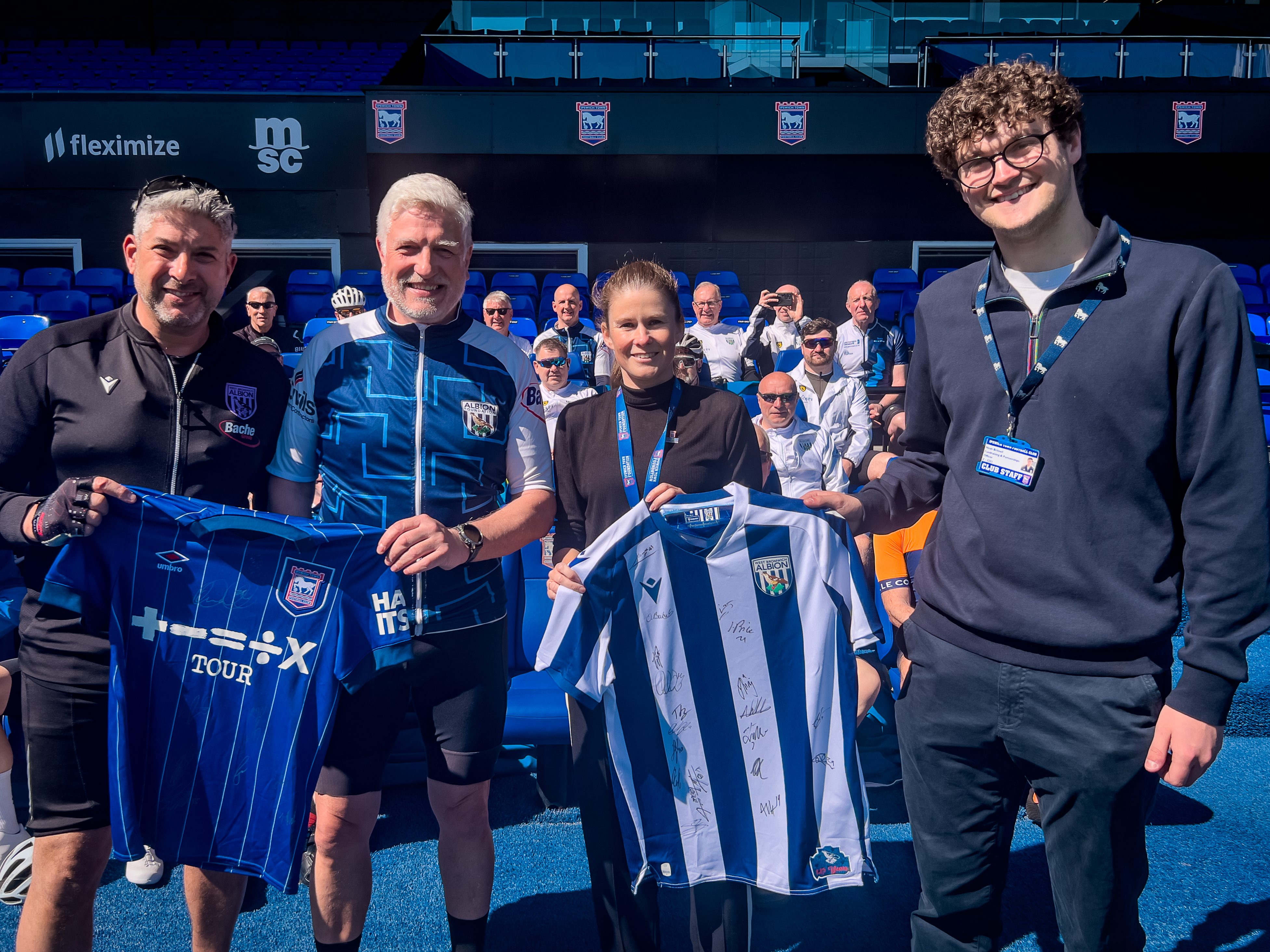 Sarah Dooley & Noah Britnell of Ipswich Town Foundation with Steve Meeson of The Albion Foundation Jude Thomson of CSCM posing with shirts for opposition teams with the cyclist in the stand.