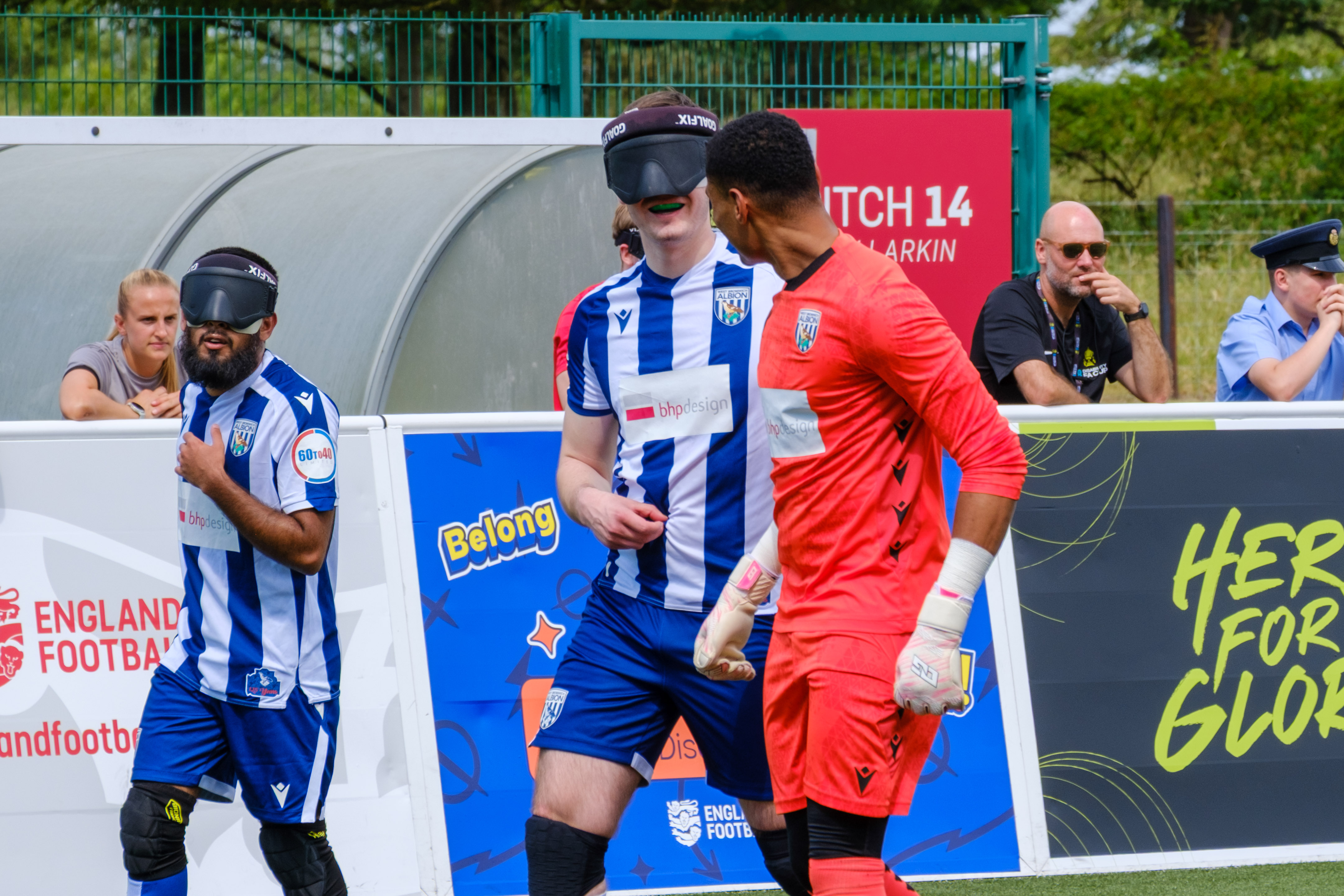 Efe Shimwell, Eesa Amjid and Owen Locke  gather near the sideline—two in blue-and-white striped kits wearing eye masks and one in a bright red goalkeeper kit—standing beside advertising boards and a team bench, with spectators and staff visible behind a pitch board.