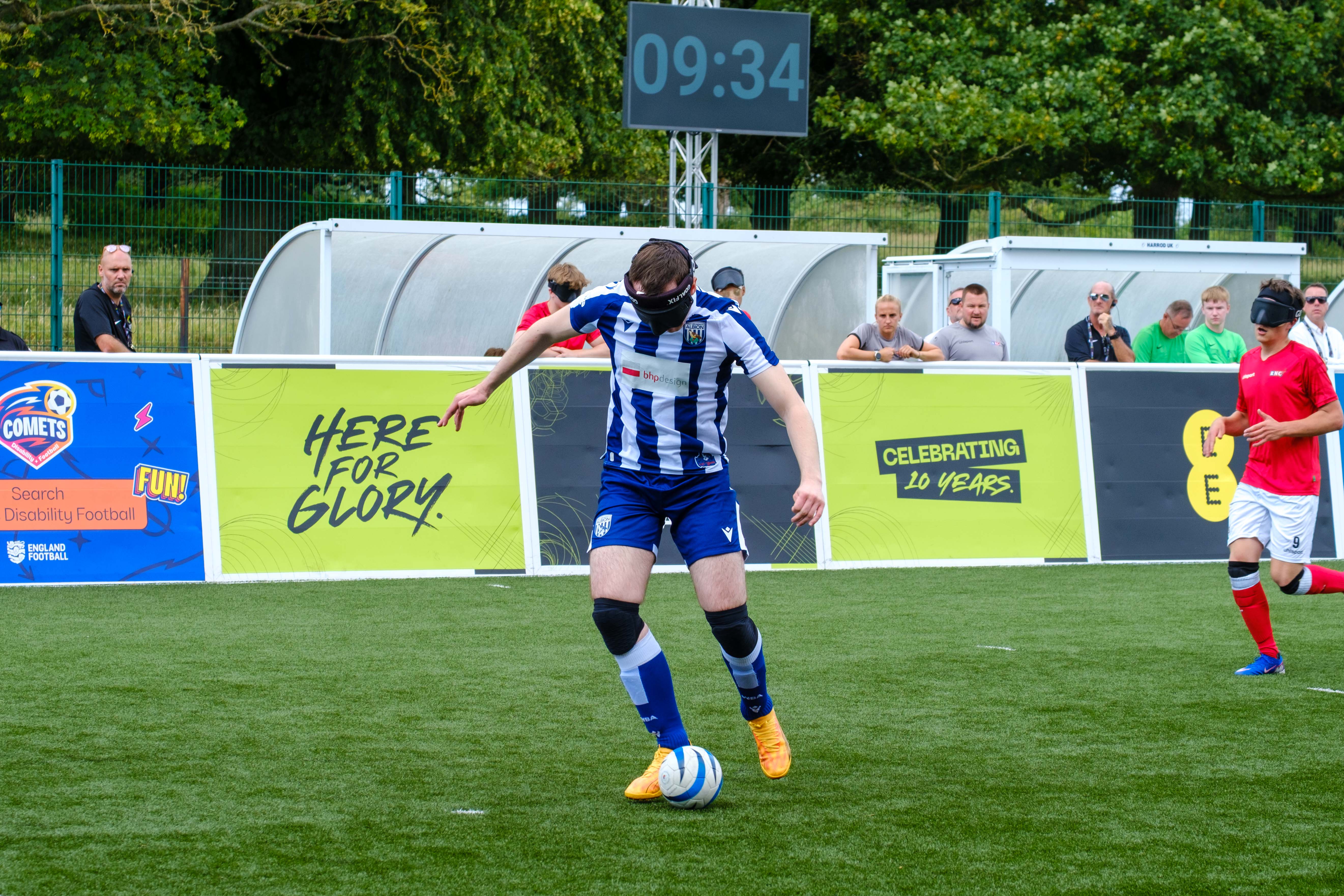 Efe Shimwell in a blue-and-white striped West Bromwich Albion kit dribbles a ball on an outdoor pitch while wearing an eye mask. A scoreboard behind shows “09:34,” and advertising boards display text including “Here for Glory” and “Celebrating 10 Years,” with spectators watching from behind the barrier.
