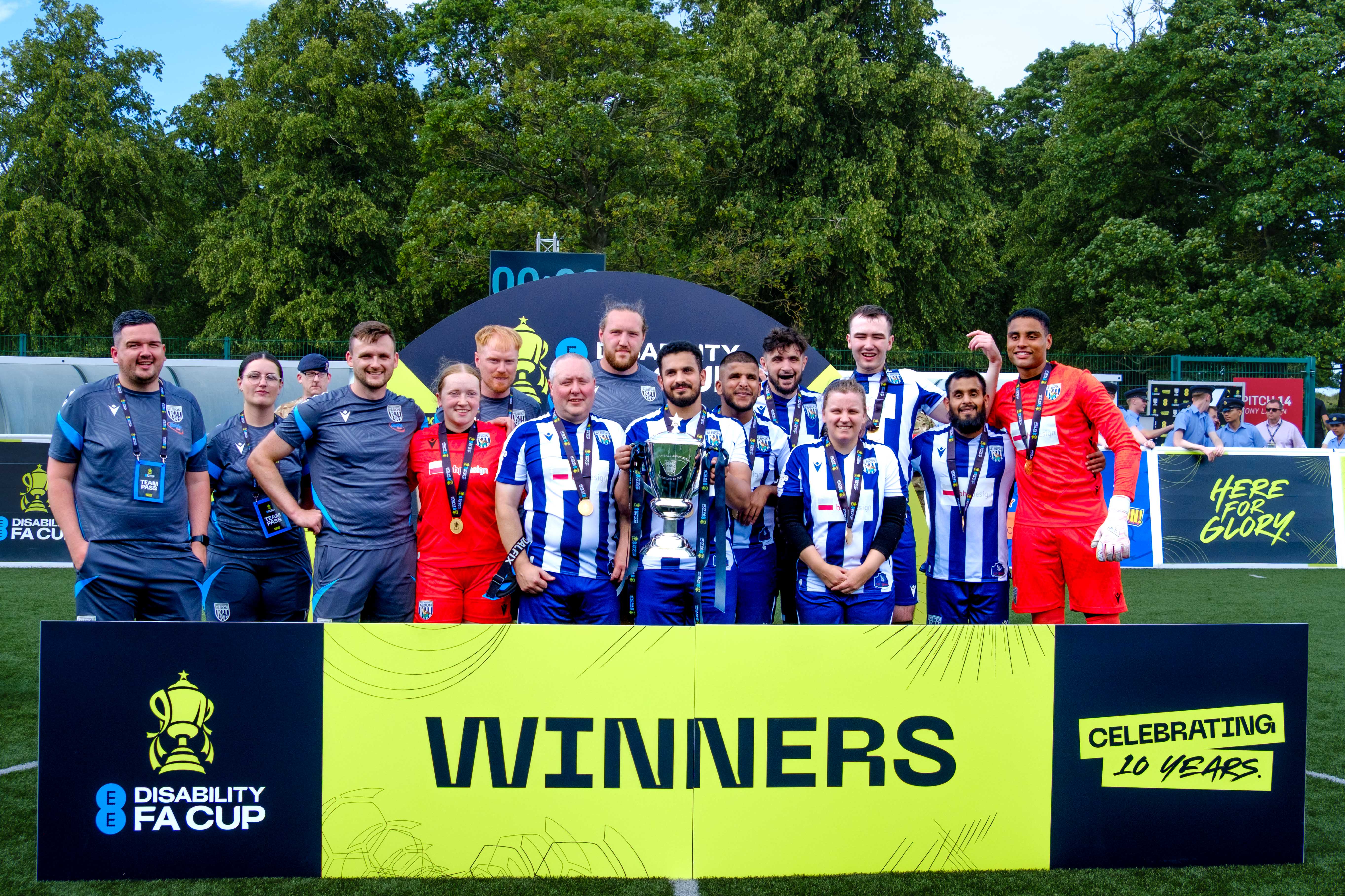 WBA Blind players on an outdoor pitch, Captain Mo Ahmed holding the trophy. A large sign in front reads “Winners” alongside the EE Disability FA Cup logo, and trees fill the background.