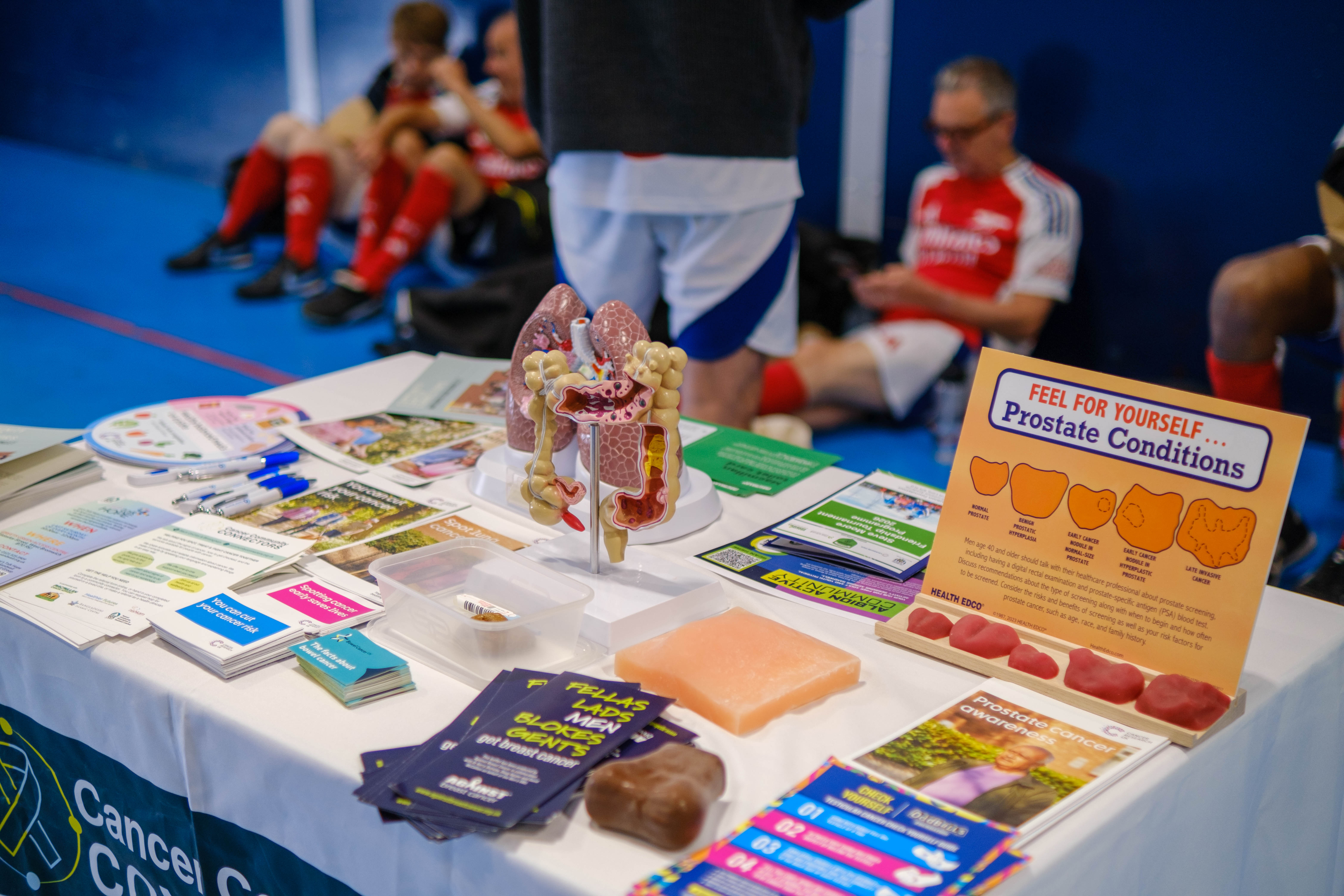 Let's Talk Hope table displaying prostate cancer awareness materials at an indoor sports venue, including anatomical models, informational leaflets.
