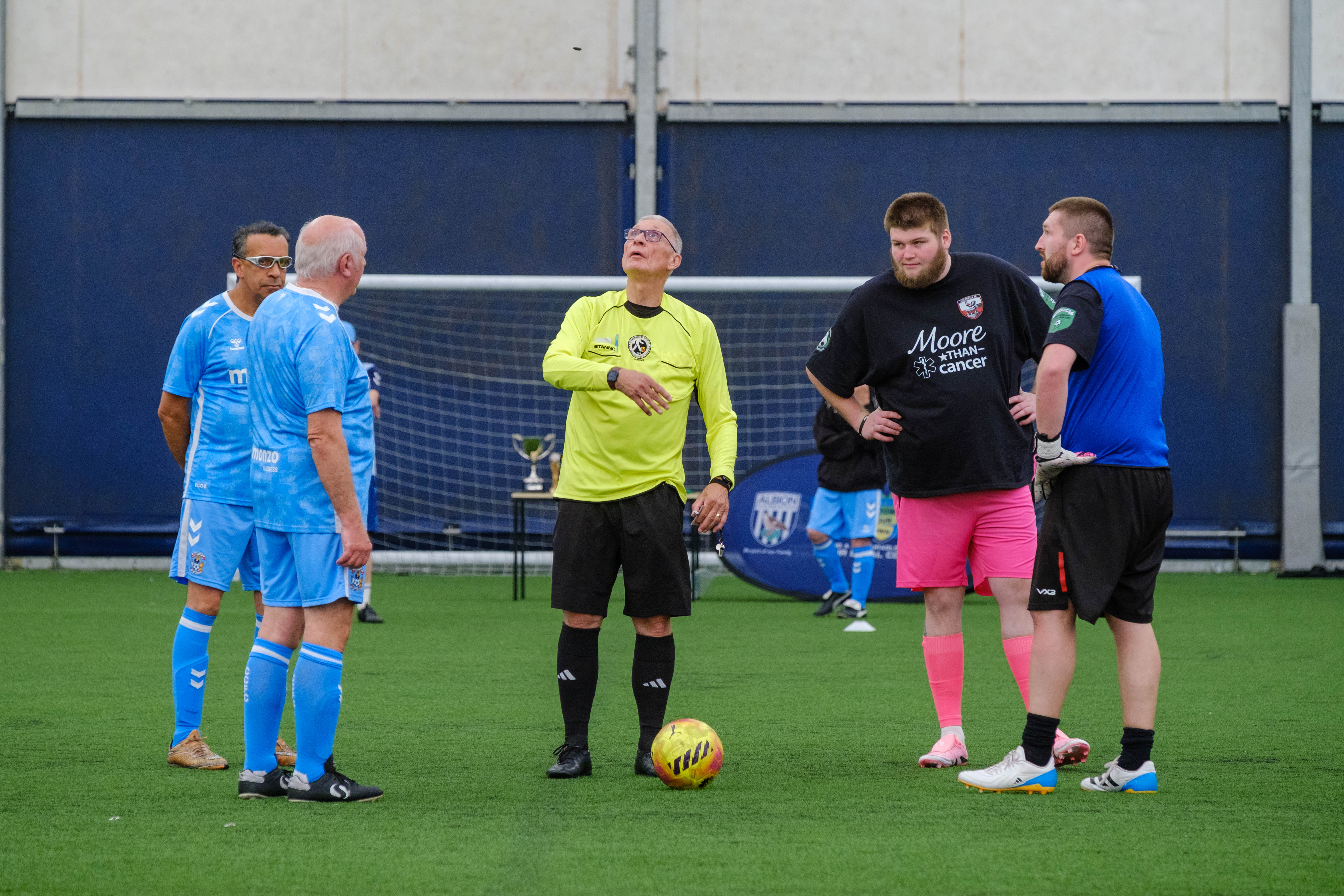 Referee in a bright yellow shirt stands on an outdoor pitch with a football at their feet, speaking to two small groups of players in blue and dark kits gathered for a pre-match coin toss, with a goal and trophy table visible behind.