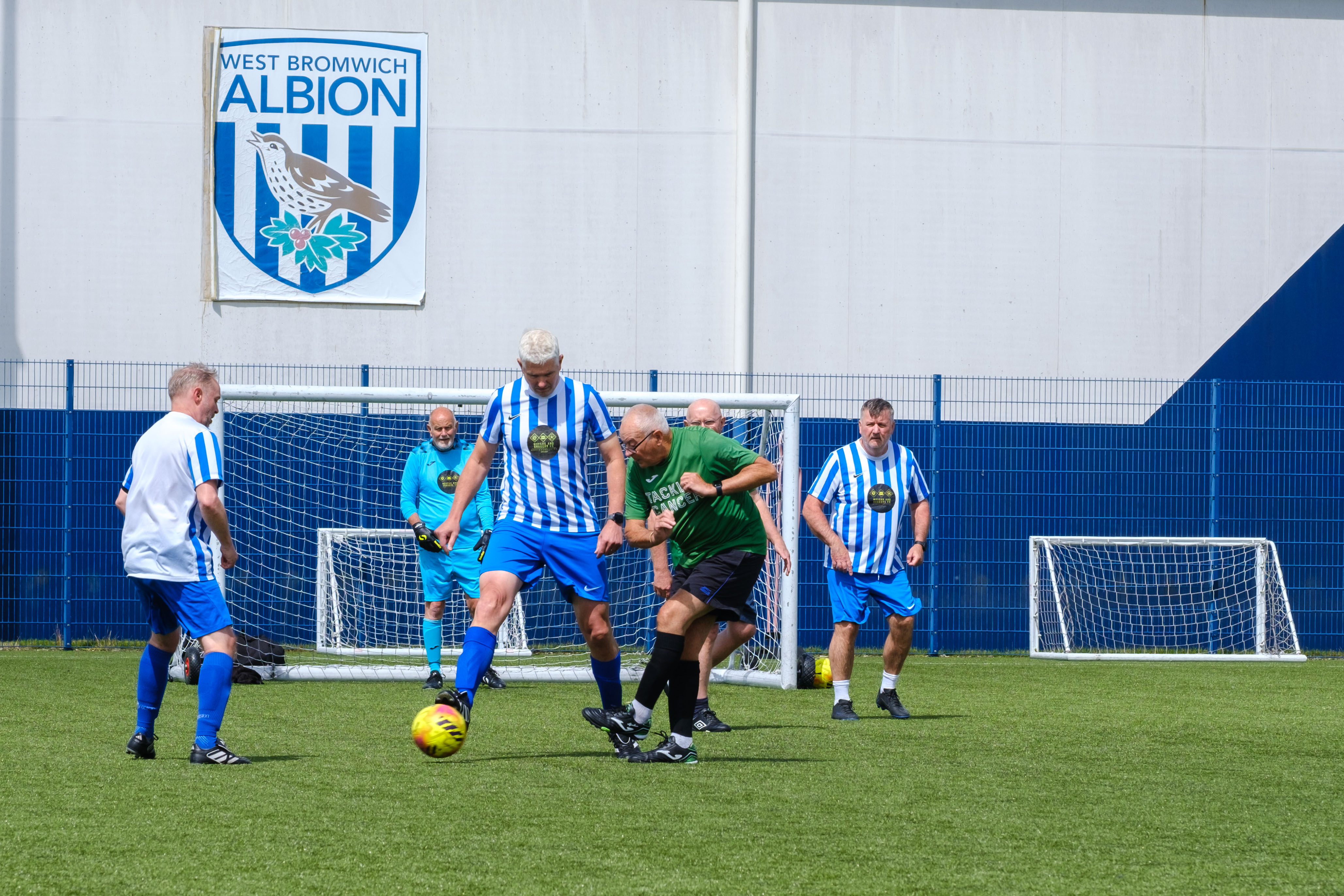 Wide shot of Big C tackling Cancer and Movers and Shakers players on the pitch with WBA logo on the dome in the background.