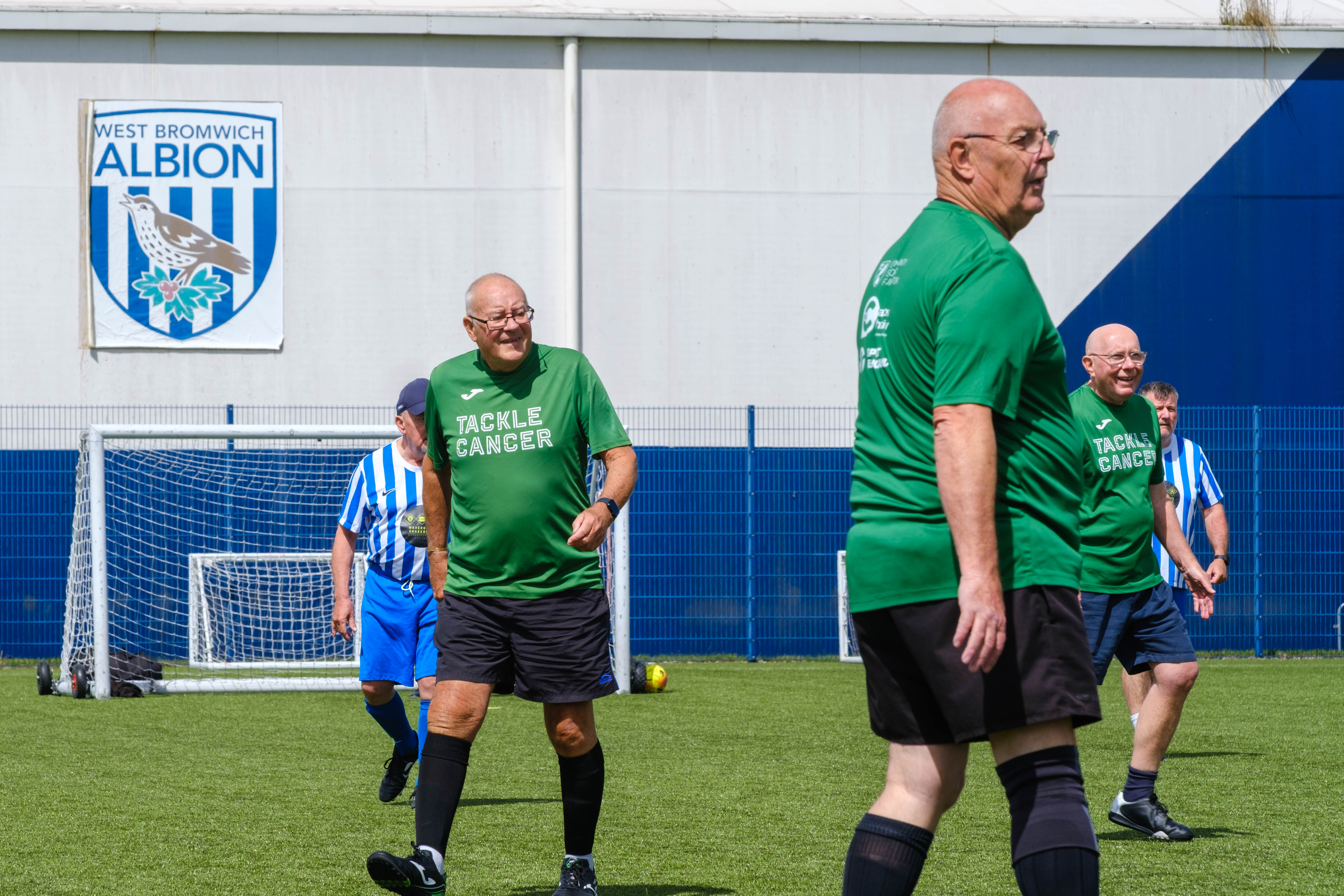 Close-up of Big C tackling Cancer players on the pitch with WBA logo on the dome in the background.