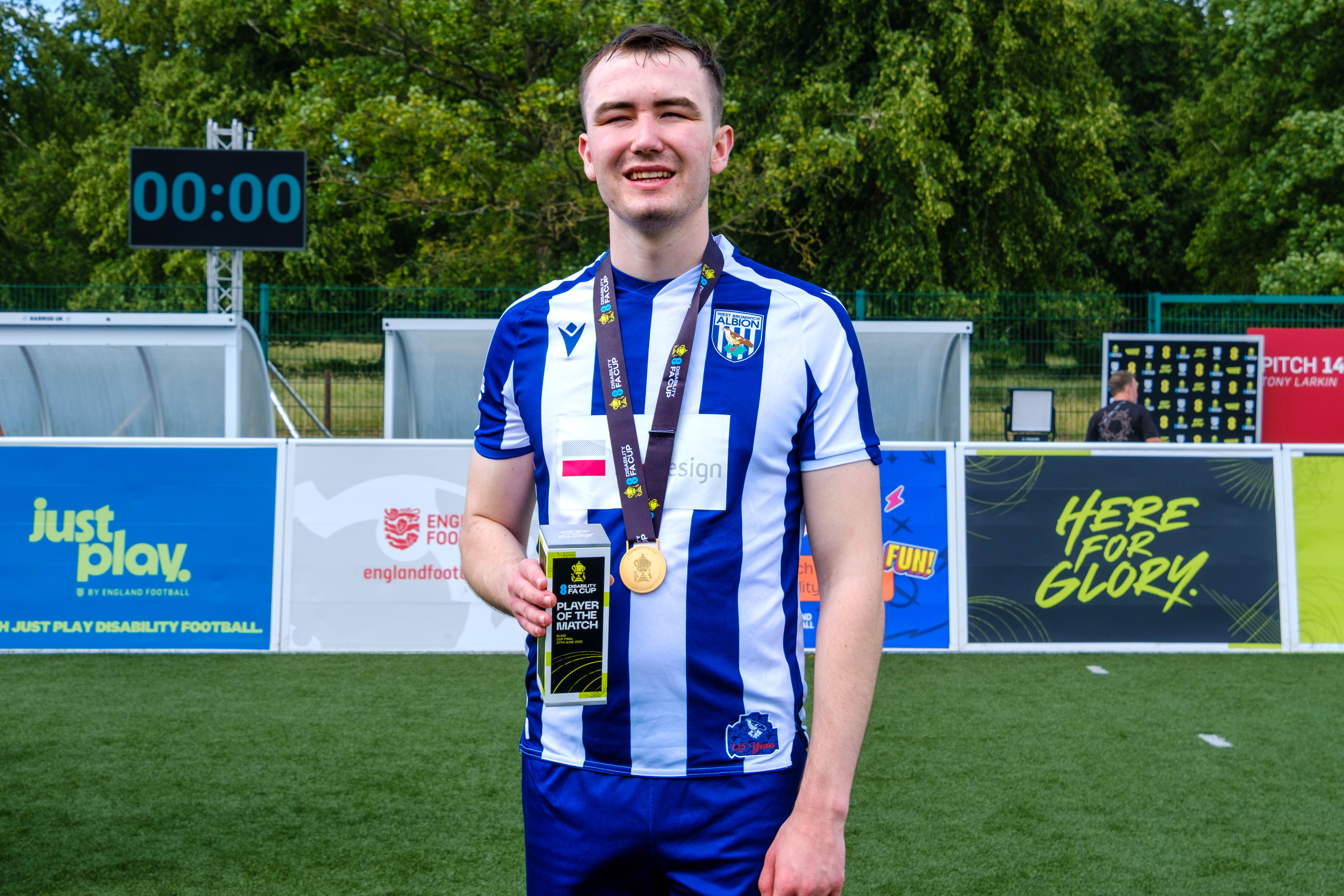 Efe Shimwell  stands on an outdoor football pitch wearing a blue-and-white striped kit, holding a Player of the Match award and wearing a medal on a ribbon around the neck. Advertising boards and a small stand are visible behind, with trees and a fence in the background.