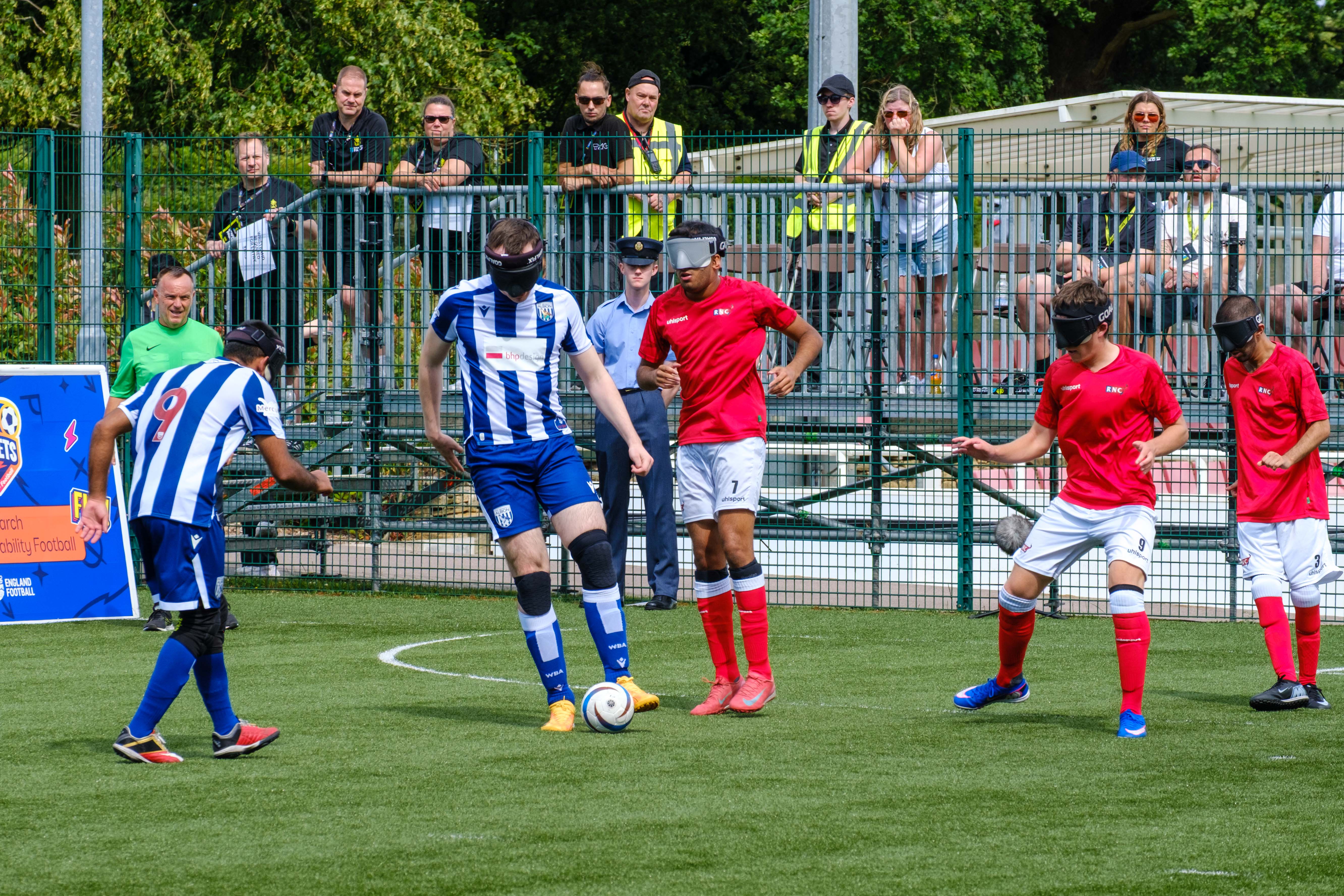Blind football players in blue‑and‑white (WBA Blind) and red (RNC) kits wear eye masks while competing on an outdoor pitch, with spectators watching behind pitch boards.