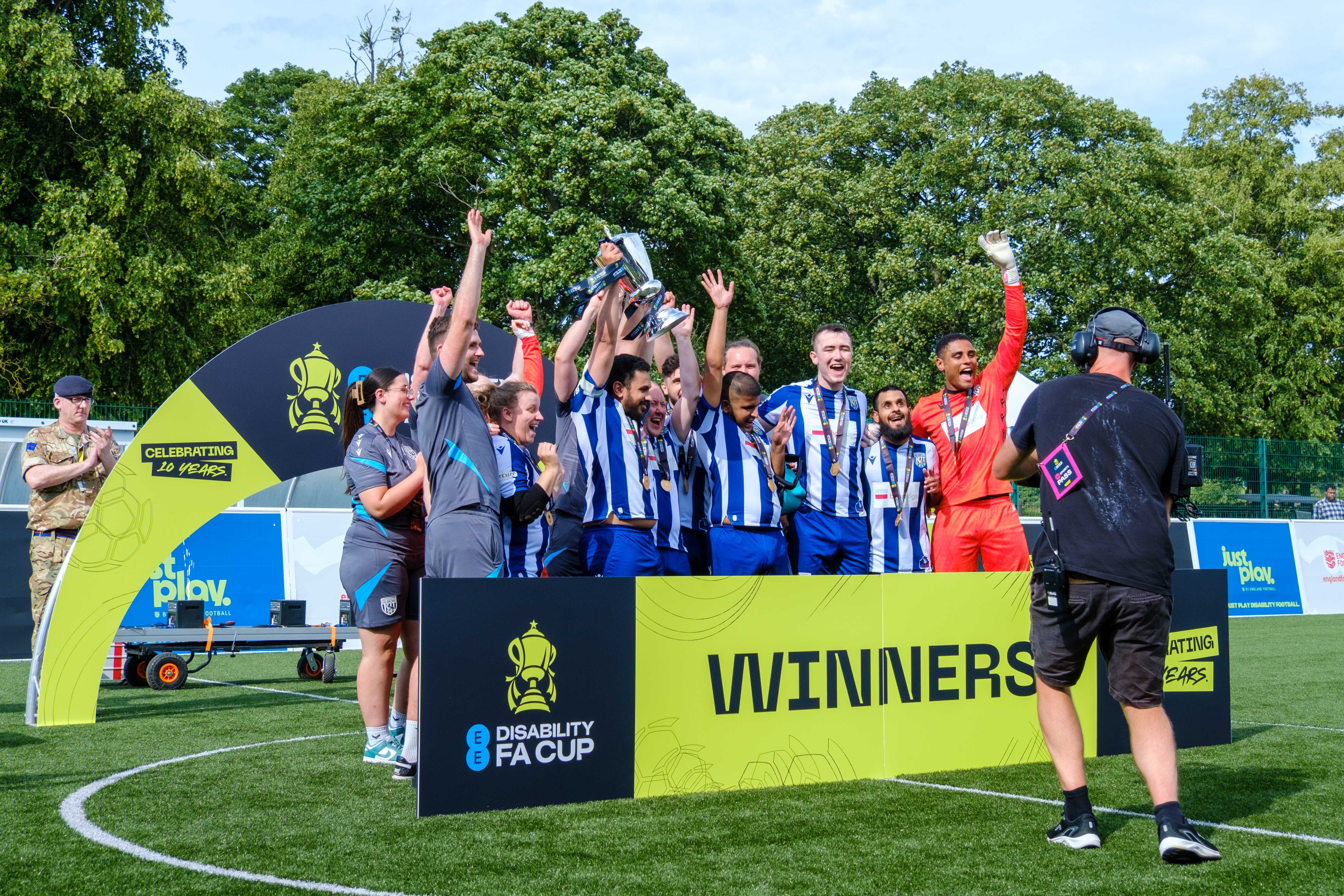 WBA Blind team on outdoor pitch, raising a trophy and their arms in celebration. A large sign in front reads “Winners” alongside the EE Disability FA Cup logo, while a camera operator films the moment and trees fill the background.