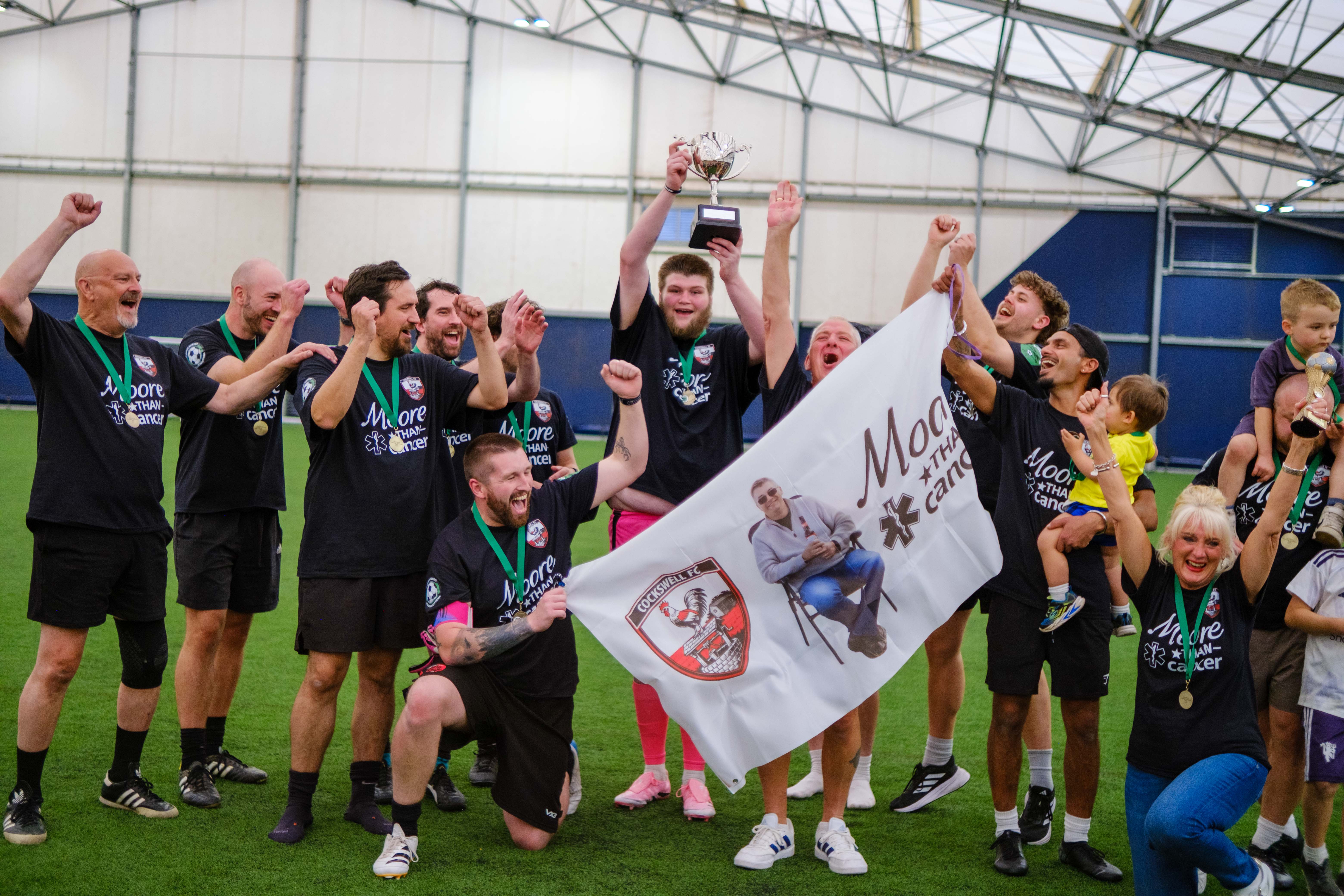 Moore than Cancer team with Ruth Moore lifting the trophy, holding a Moore than Cancer tribute banner.
