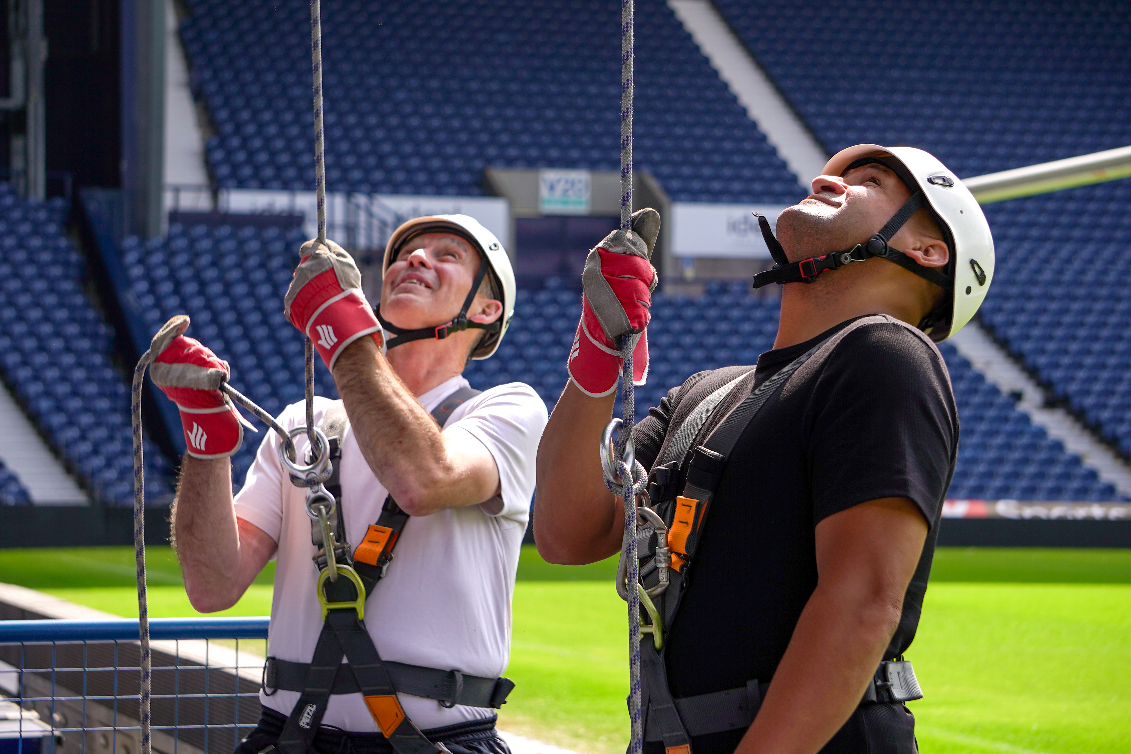 Super Bob Taylor and Nigel Quashie wearing helmets, harnesses and safety gloves prepare for an abseil at The Hawthorns, looking up at the ropes with the stadium seating visible behind them.