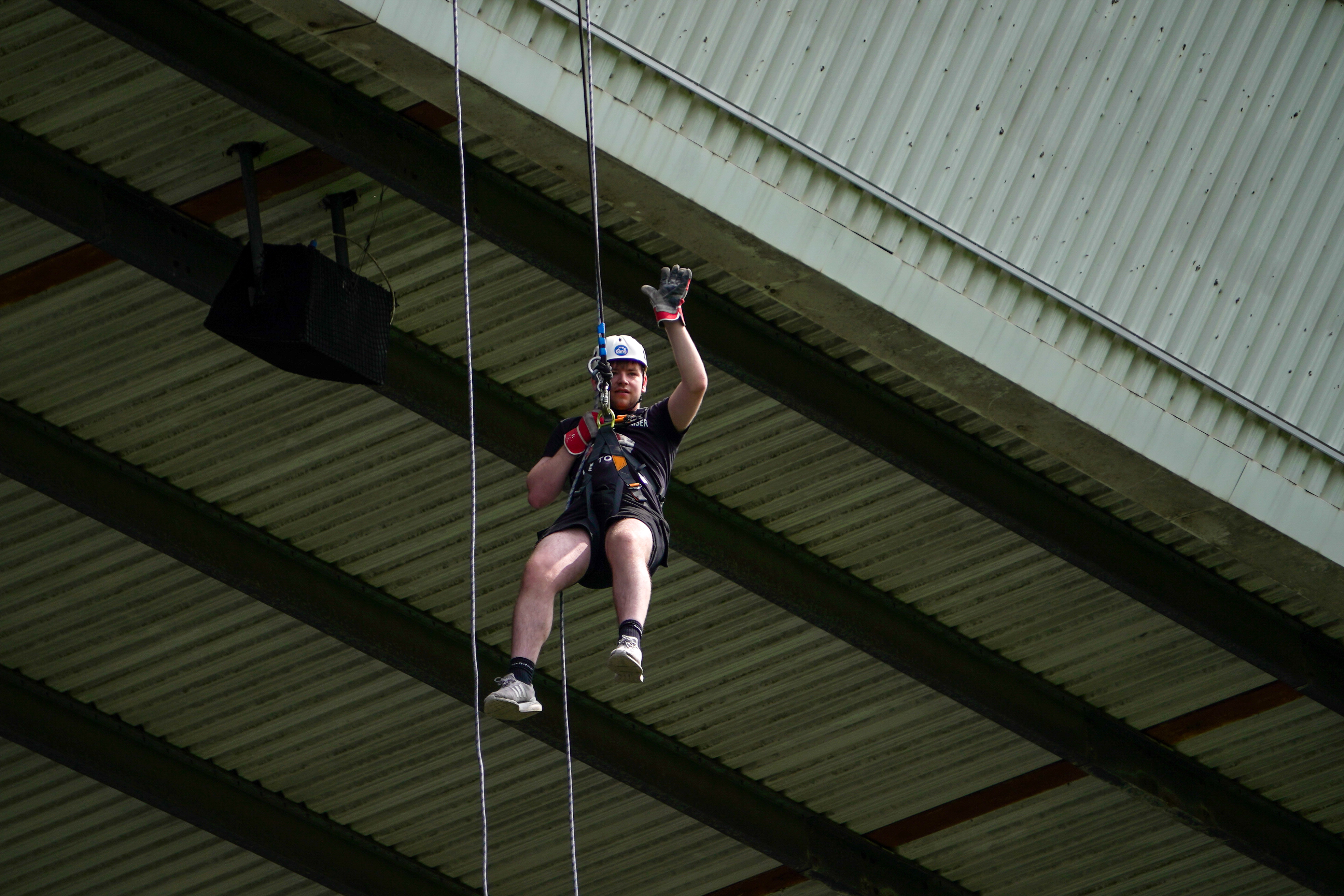 Participant wearing a helmet and harness waves while abseiling beneath the stadium roof at The Hawthorns.