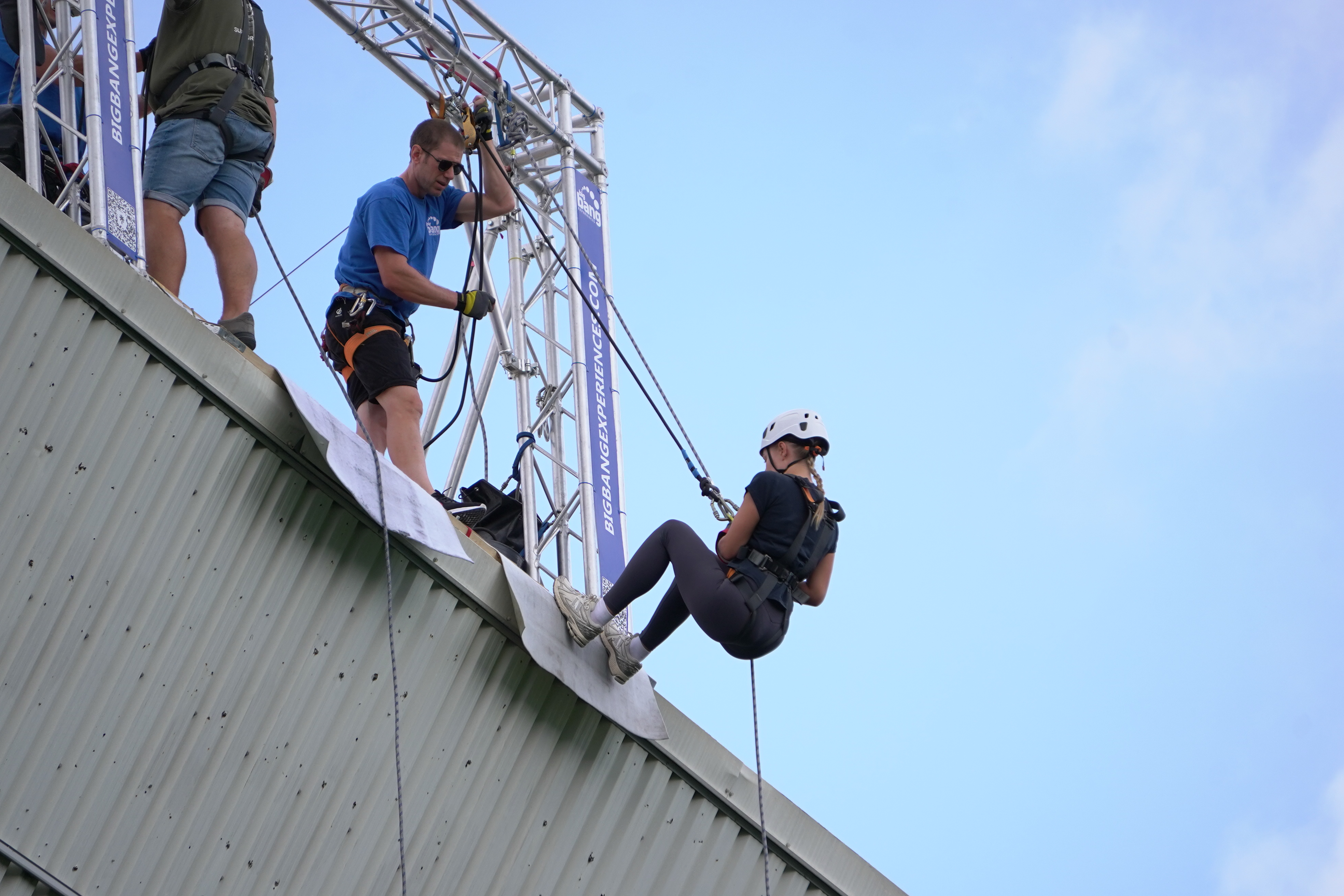 Participant wearing a helmet and safety harness begins an abseil from the roof of a stadium, guided by an instructor standing beside the rope system under a clear blue sky.