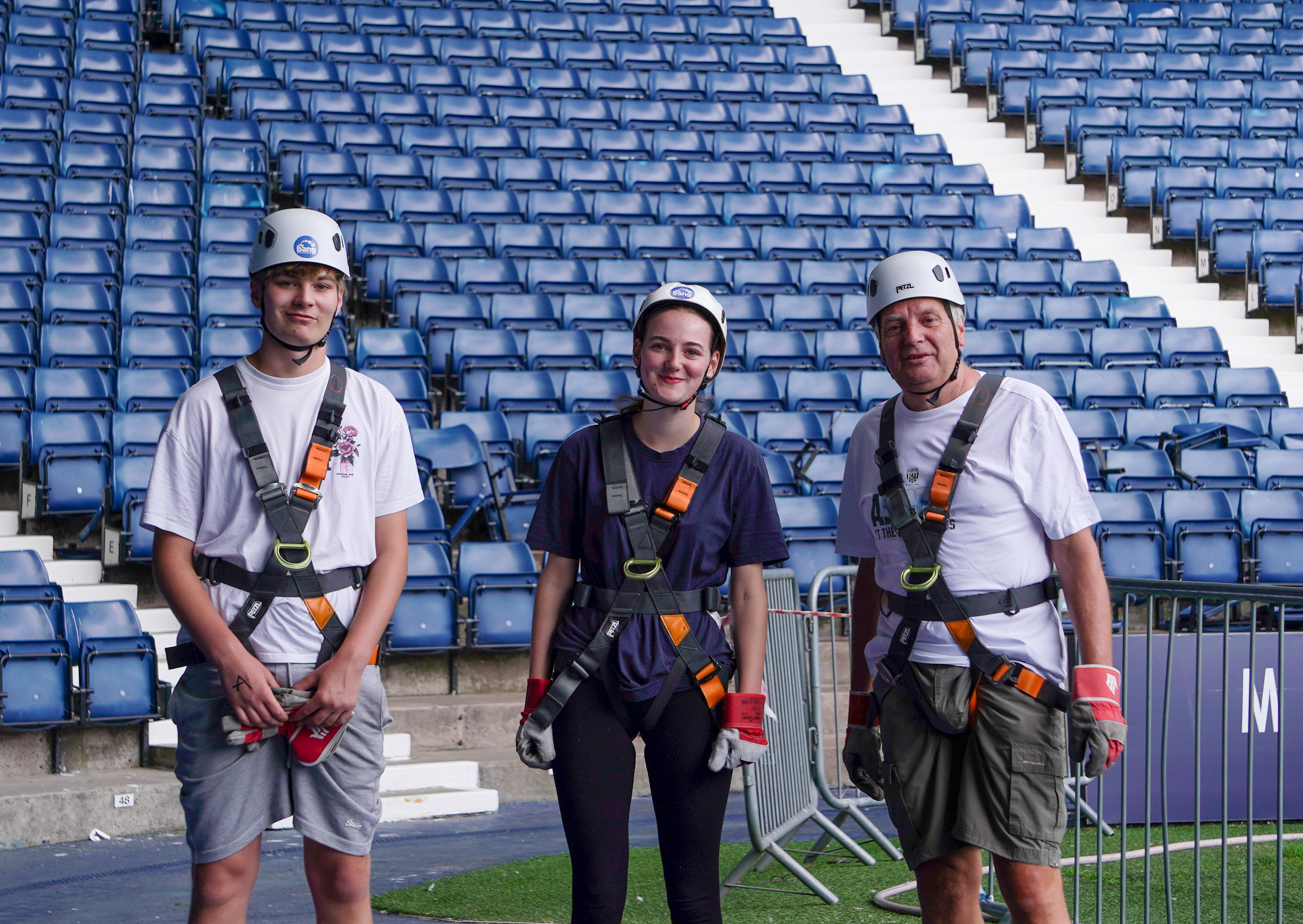 Three participants wearing helmets and safety harnesses pose for a photo on the pitchside area at The Hawthorns after completing an abseil.