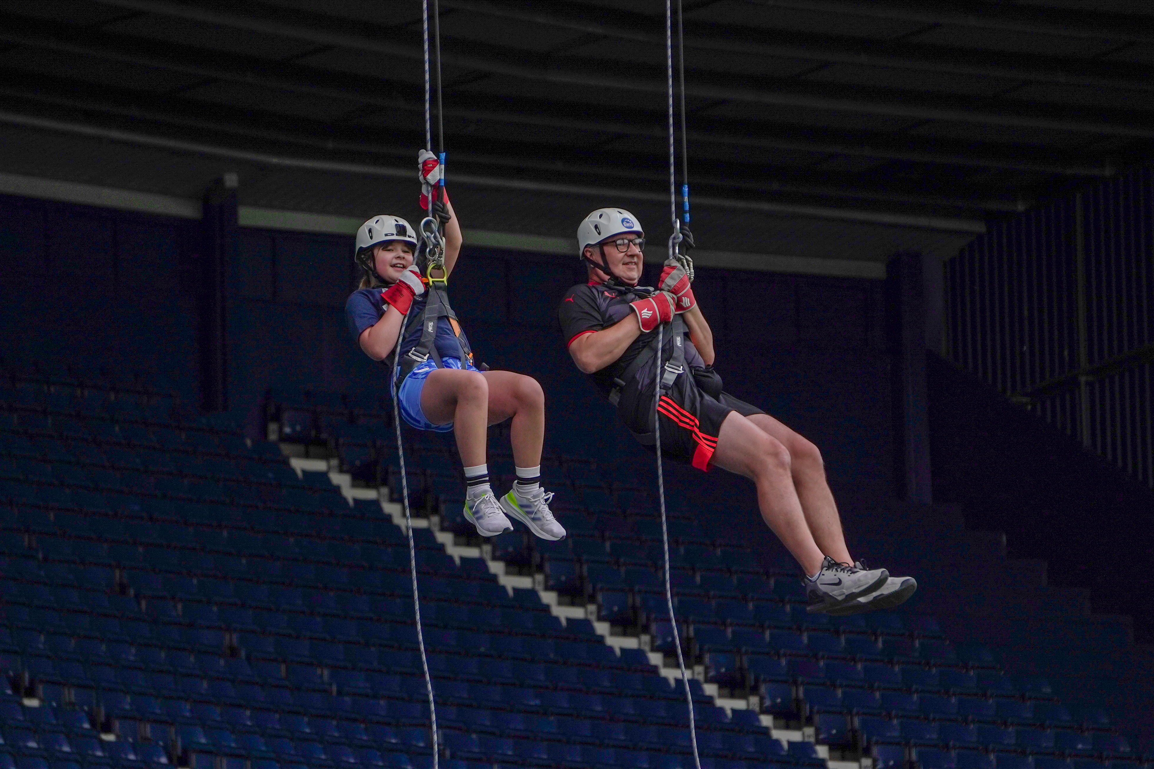 Two participants wearing helmets and safety harnesses descend on ropes inside a sports arena, suspended high above rows of blue stadium seats during a supervised abseiling activity.