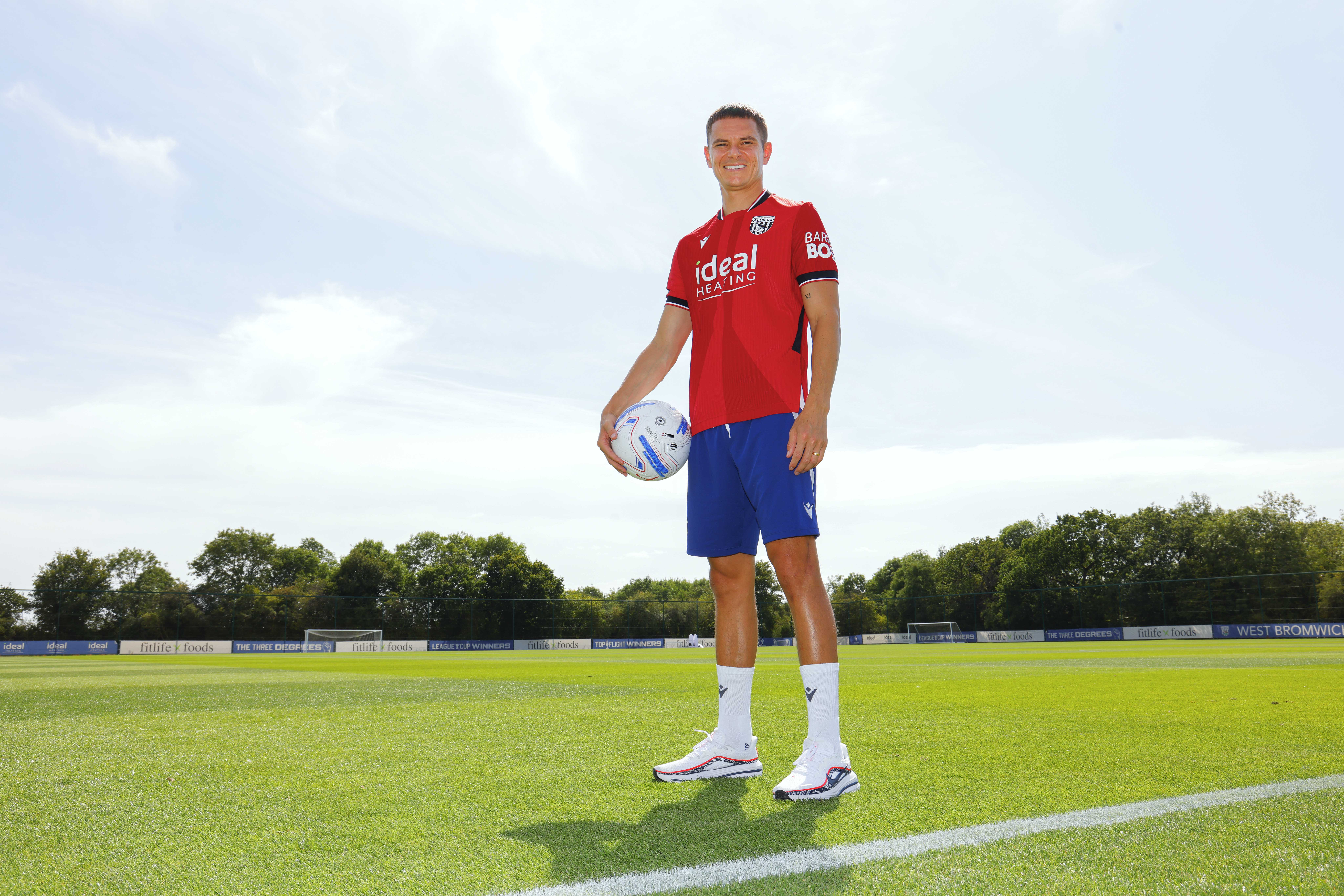 Conor Townsend smiling and looking at the camera while stood out on the training pitch in the red away kit with a ball in his hand 