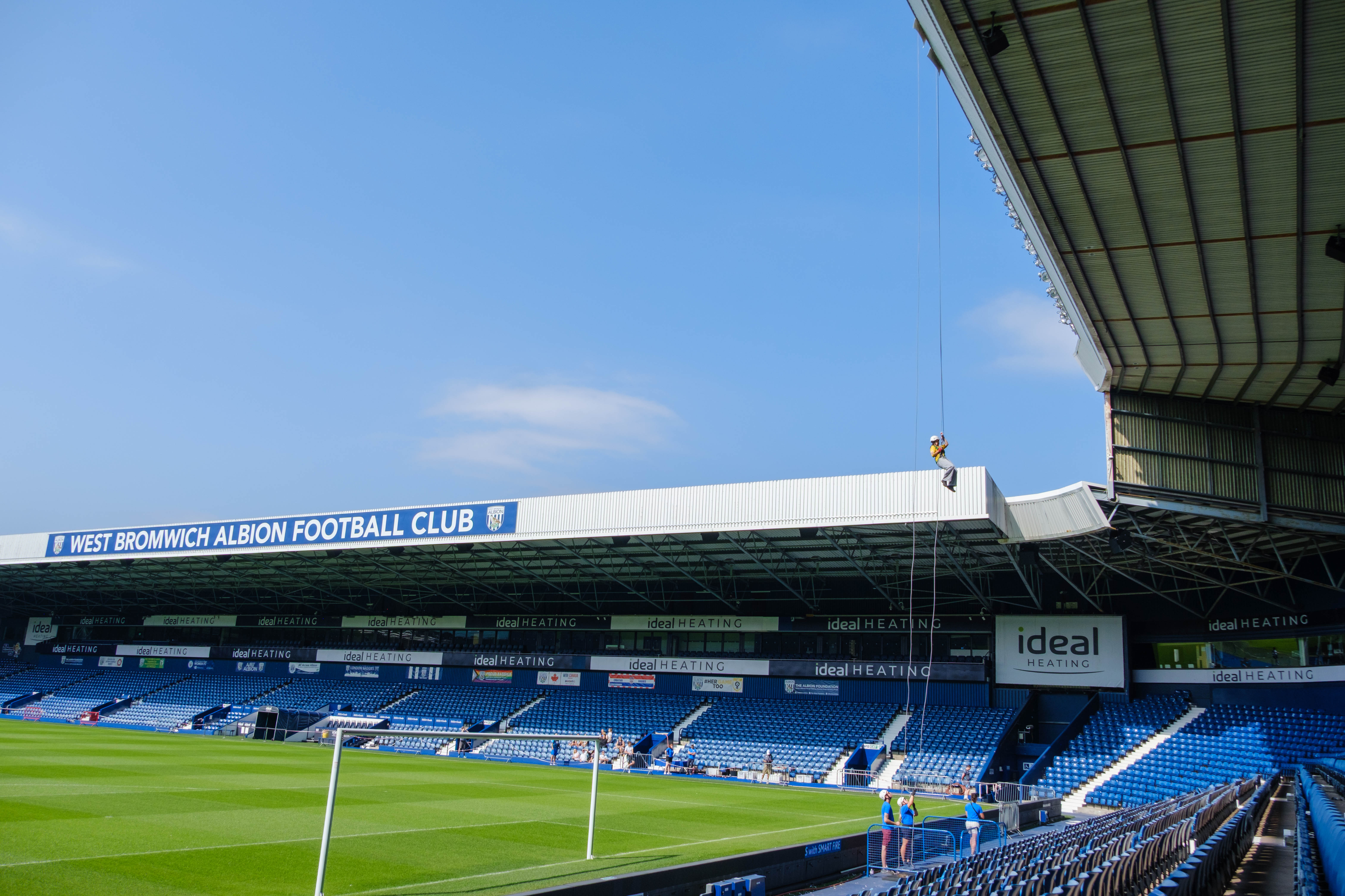 Participant abseils from the roof of The Hawthorns high above the West Bromwich Albion pitch, with blue seating and stadium stands visible below.