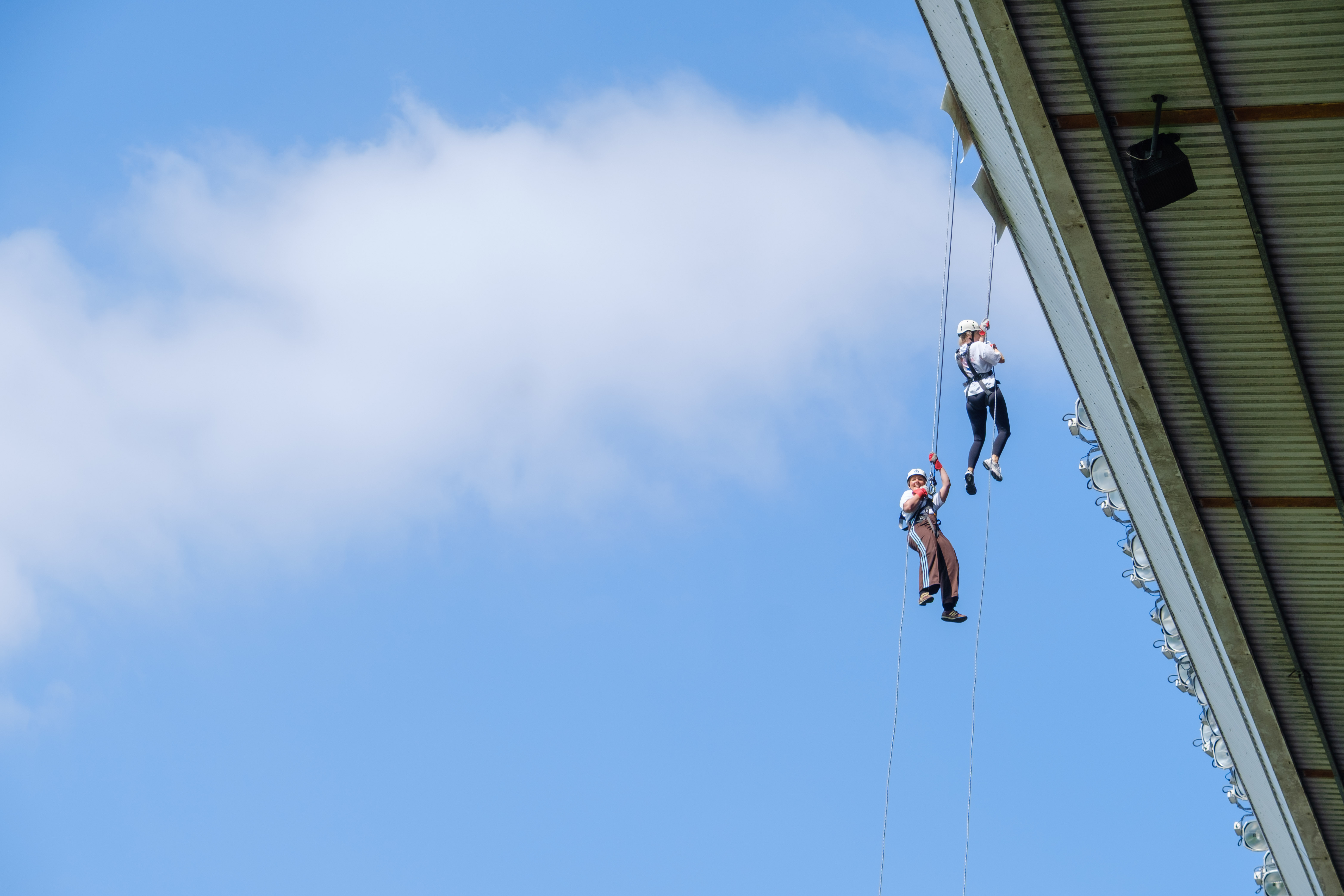 Two participants abseil side by side from the roof of The Hawthorns, suspended high above the stadium against a blue sky.