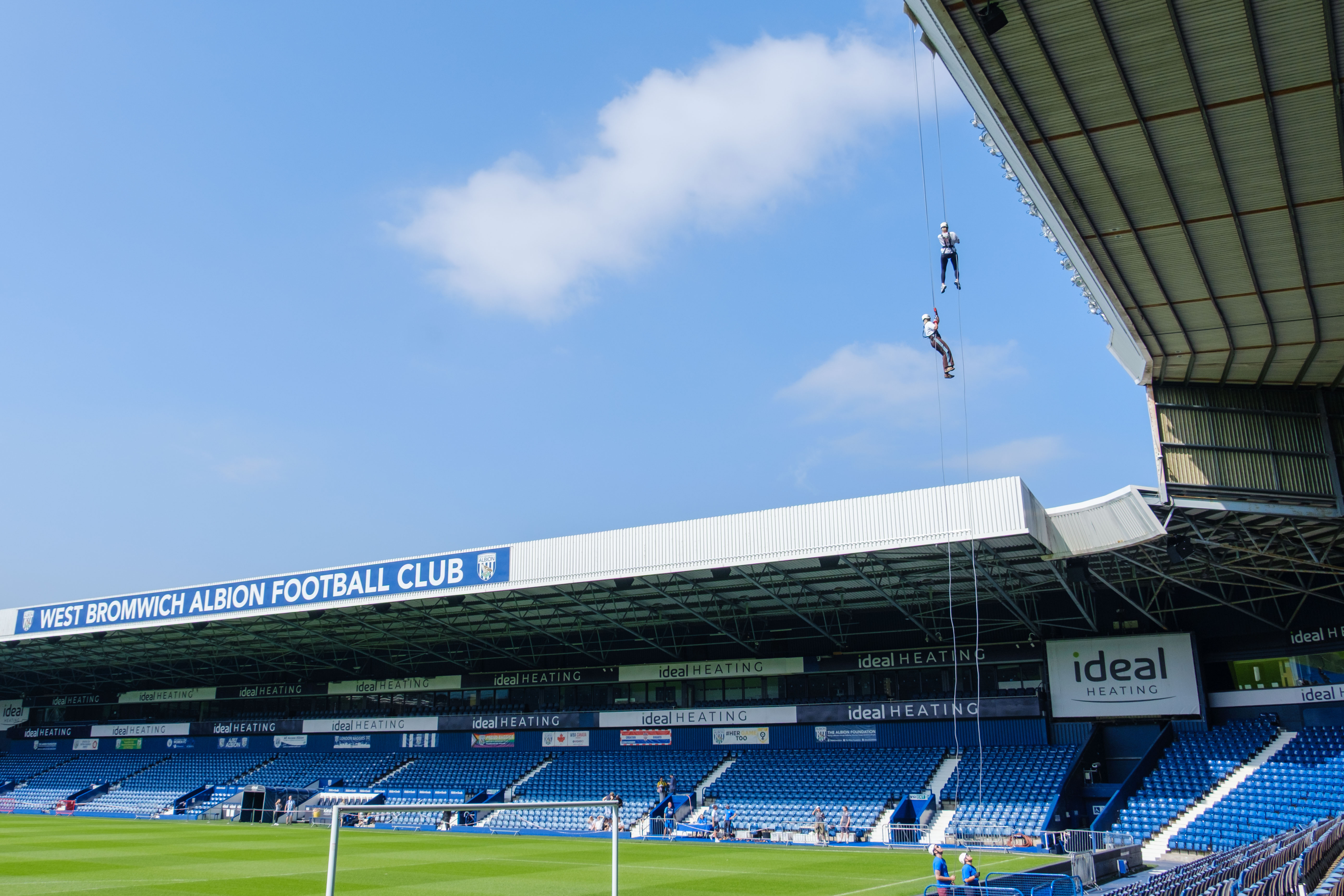 Two participants abseil from the roof of The Hawthorns on ropes suspended high above the pitch, with the West Bromwich Albion Football Club stand and rows of blue seats visible below under a clear sky.