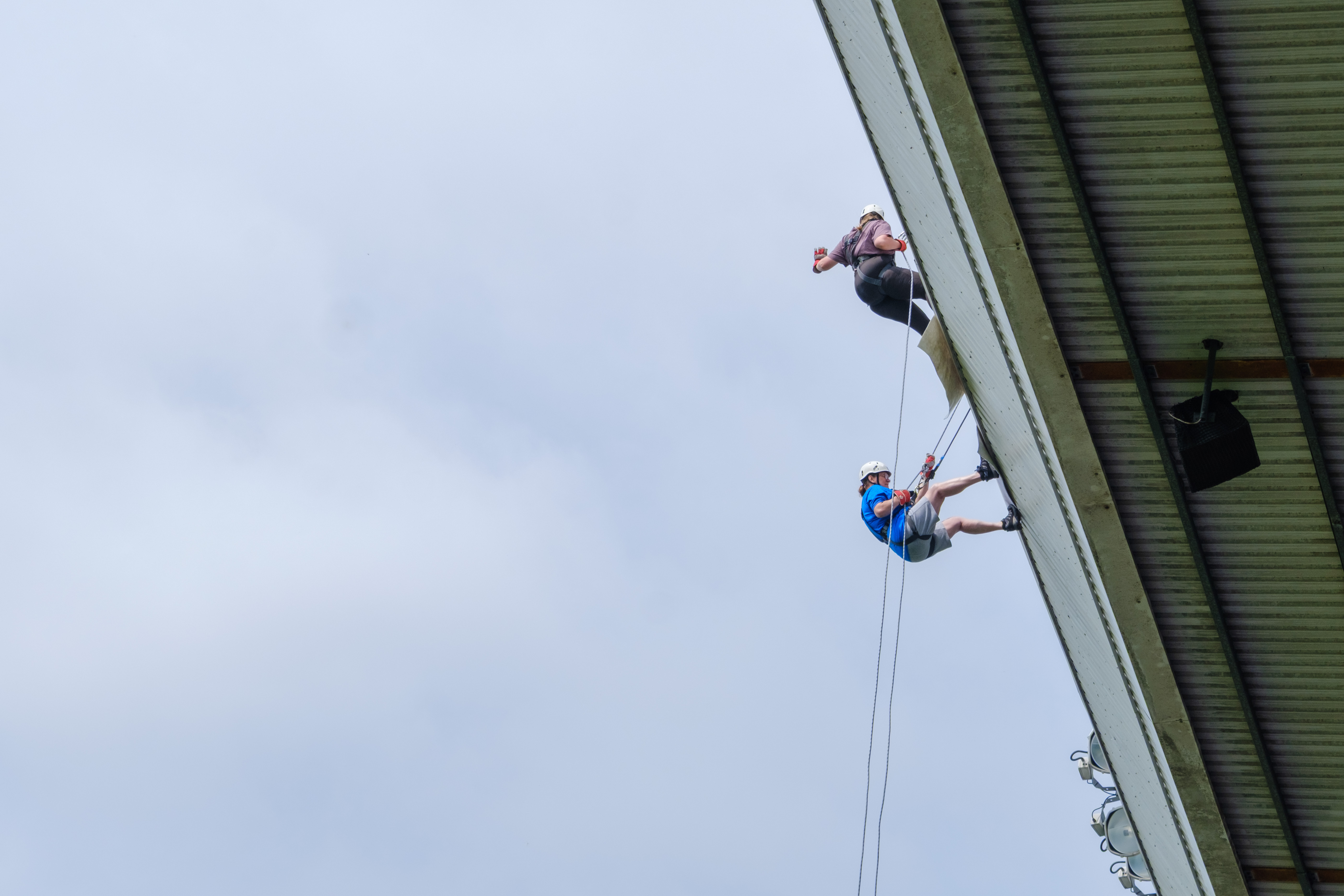Two participants abseil side by side from the roof of The Hawthorns, suspended on ropes against the stadium structure and sky.