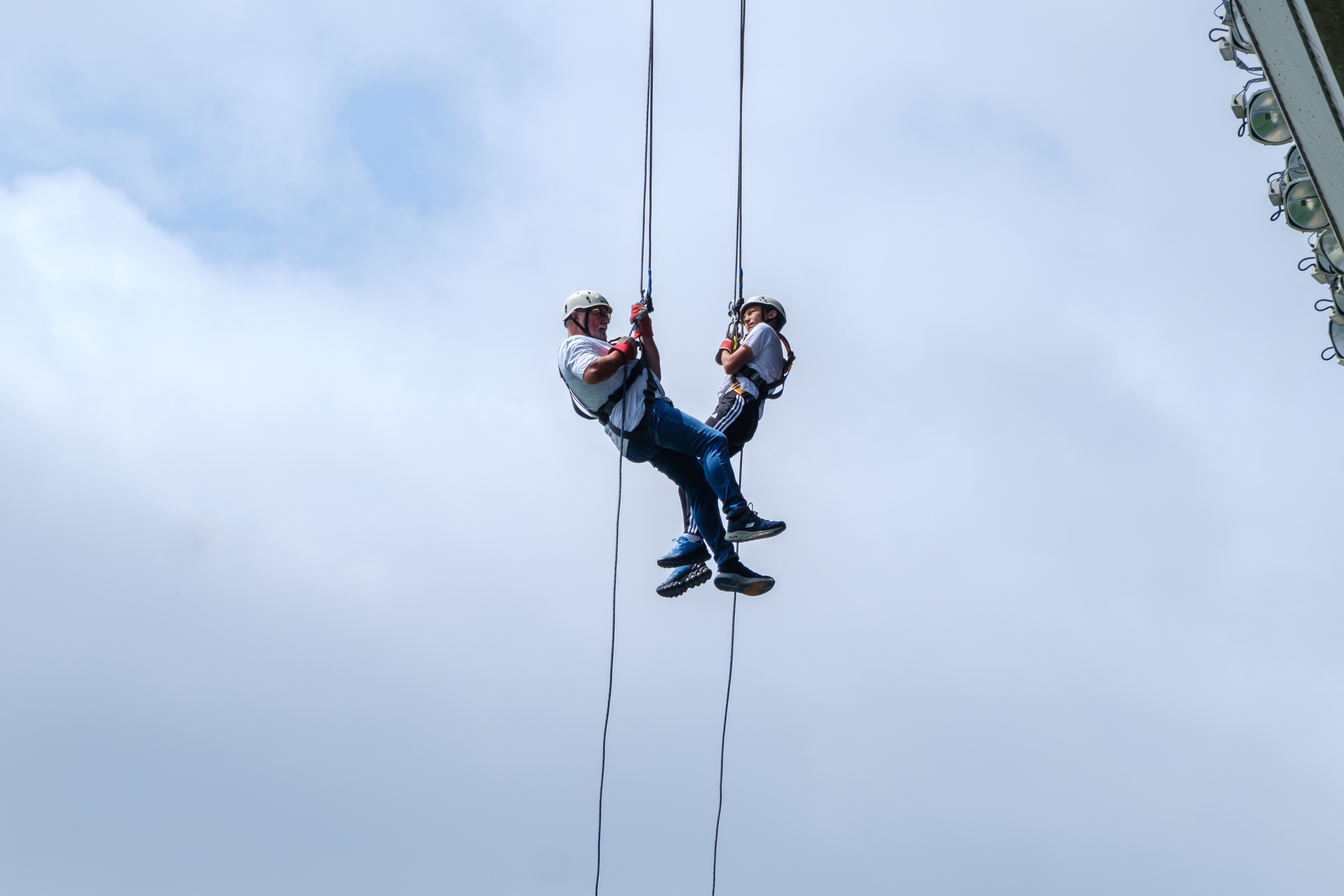 Two participants descend on ropes during a tandem abseil from the roof of The Hawthorns, suspended against a cloudy sky.