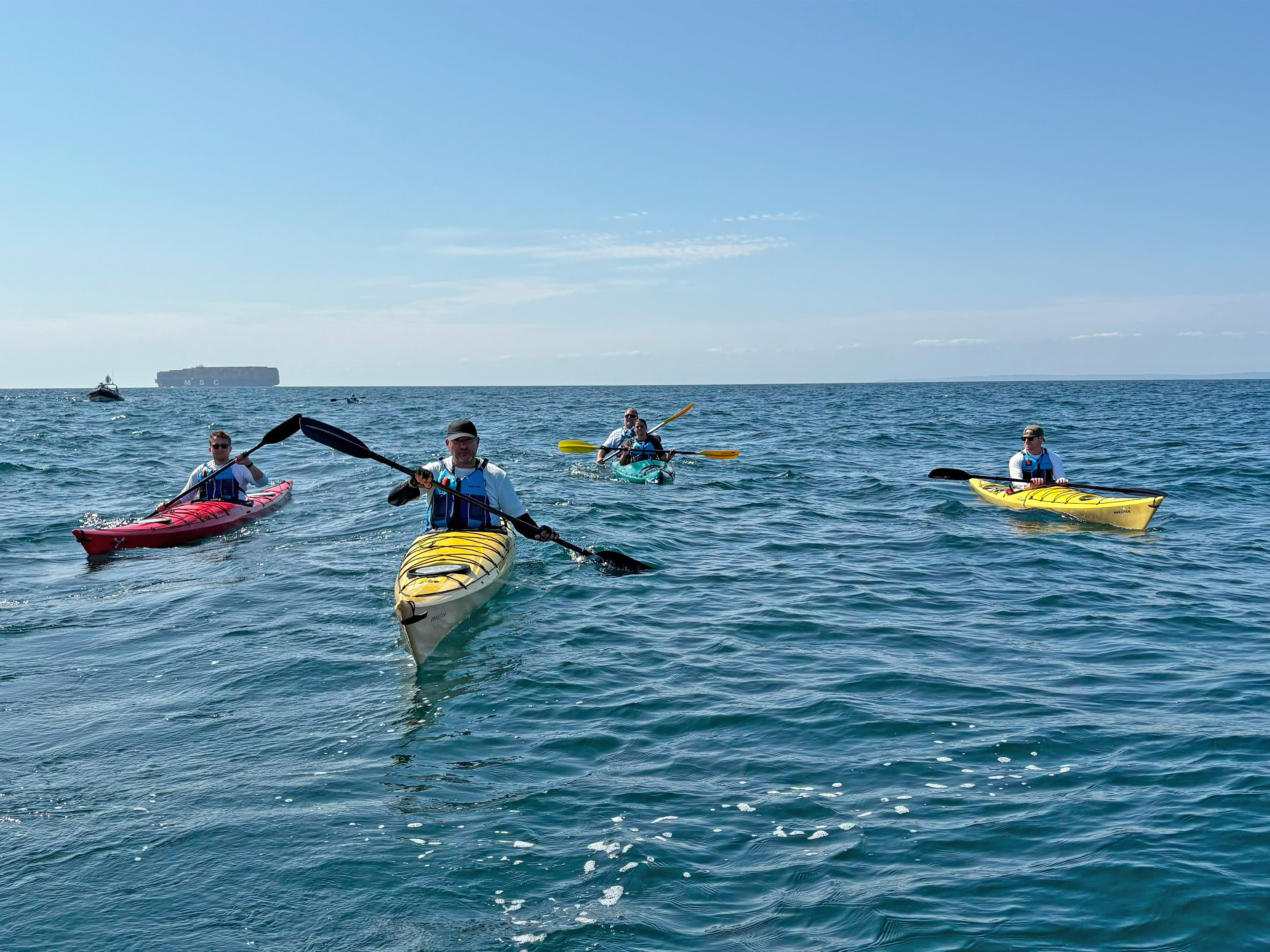 People kayaking on open water in brightly coloured kayaks, paddling in a group under clear blue skies, with a large cargo ship visible on the horizon.
