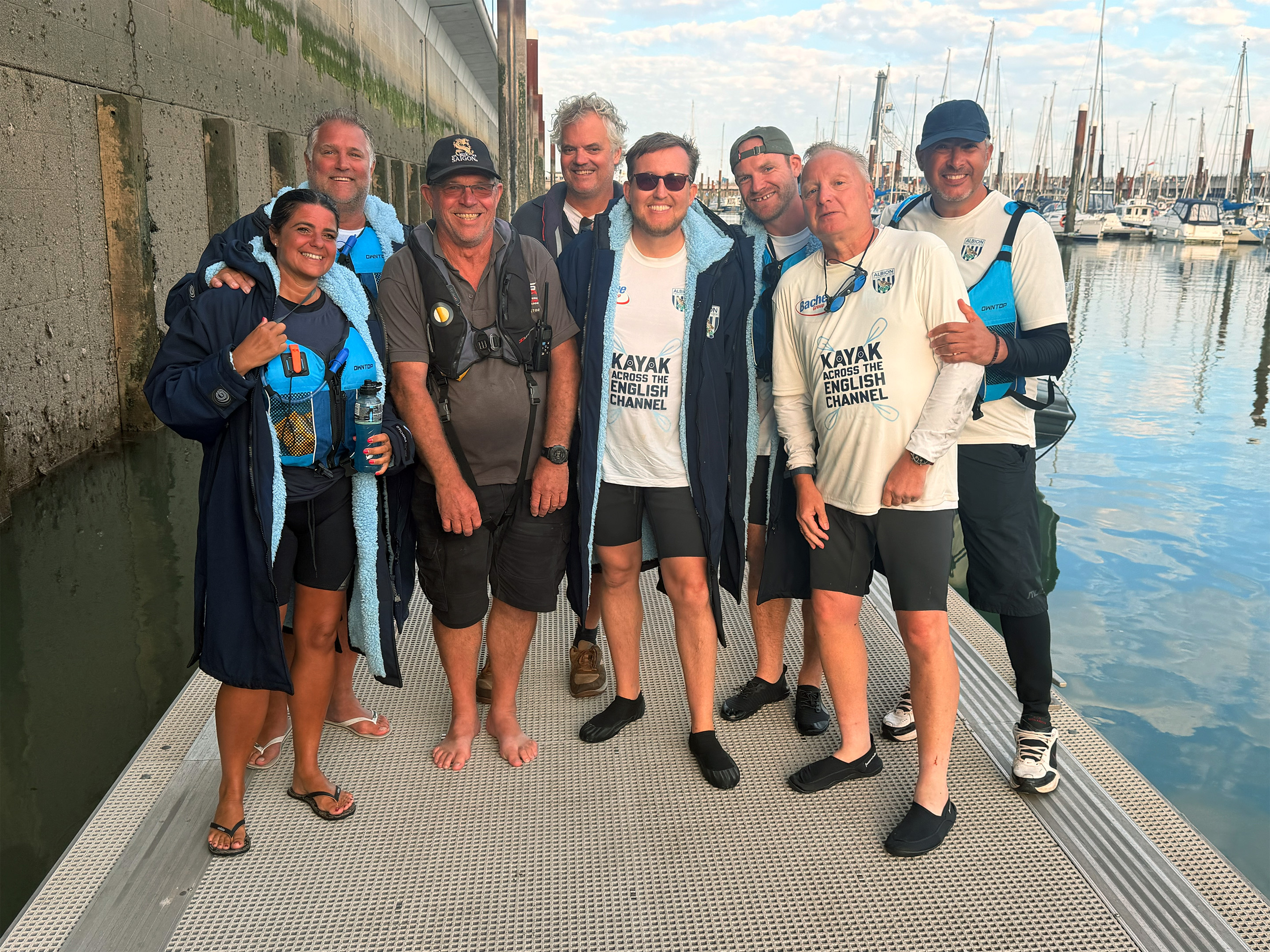 Group photo of participants and support crew standing on a marina pontoon after completing an English Channel kayaking challenge.