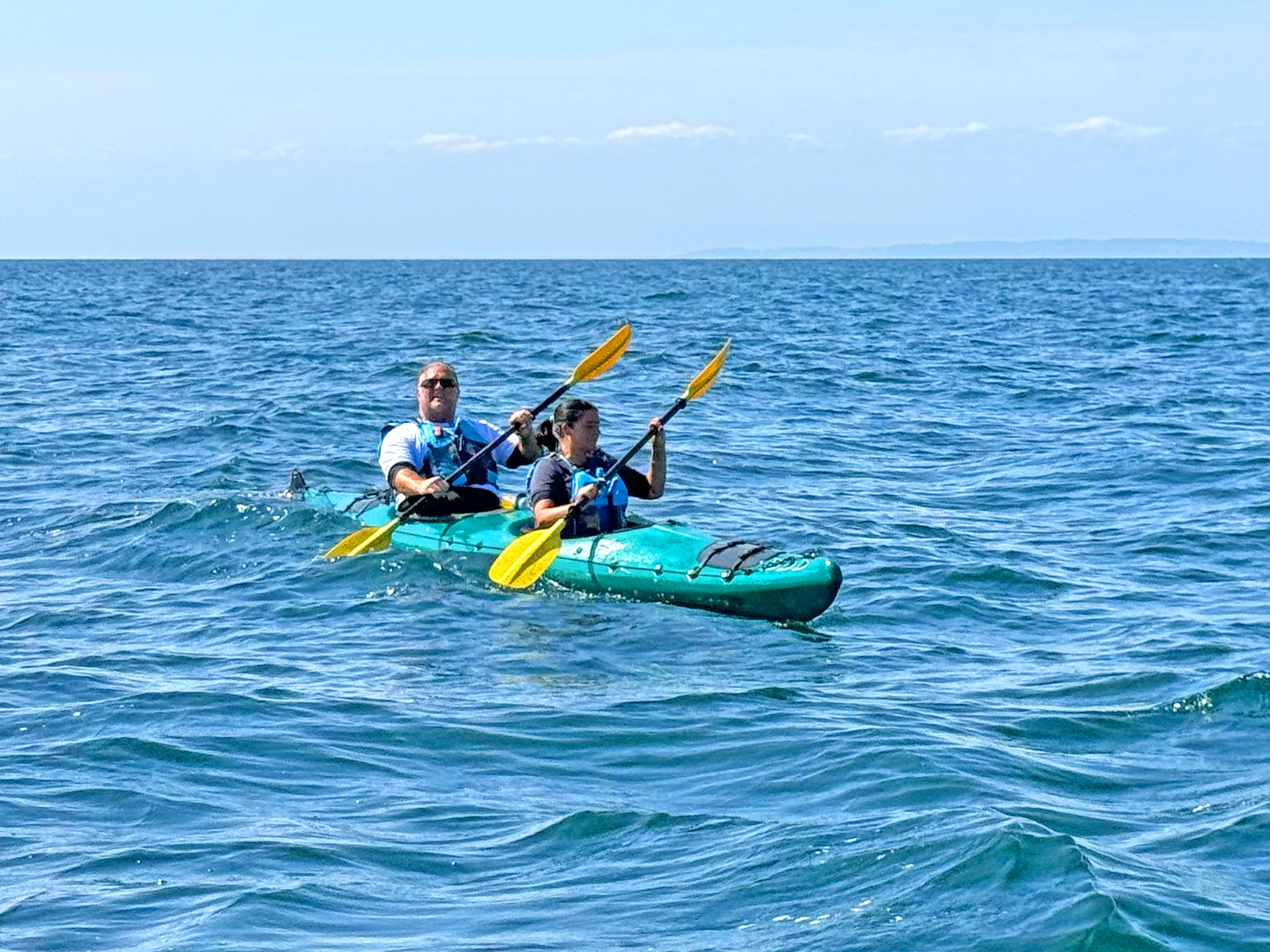 Andy Johnson & Kelly paddle a tandem kayak across open water during an English Channel kayaking challenge, wearing buoyancy aids as they navigate small waves under a clear sky.Provide your feedback on BizChat