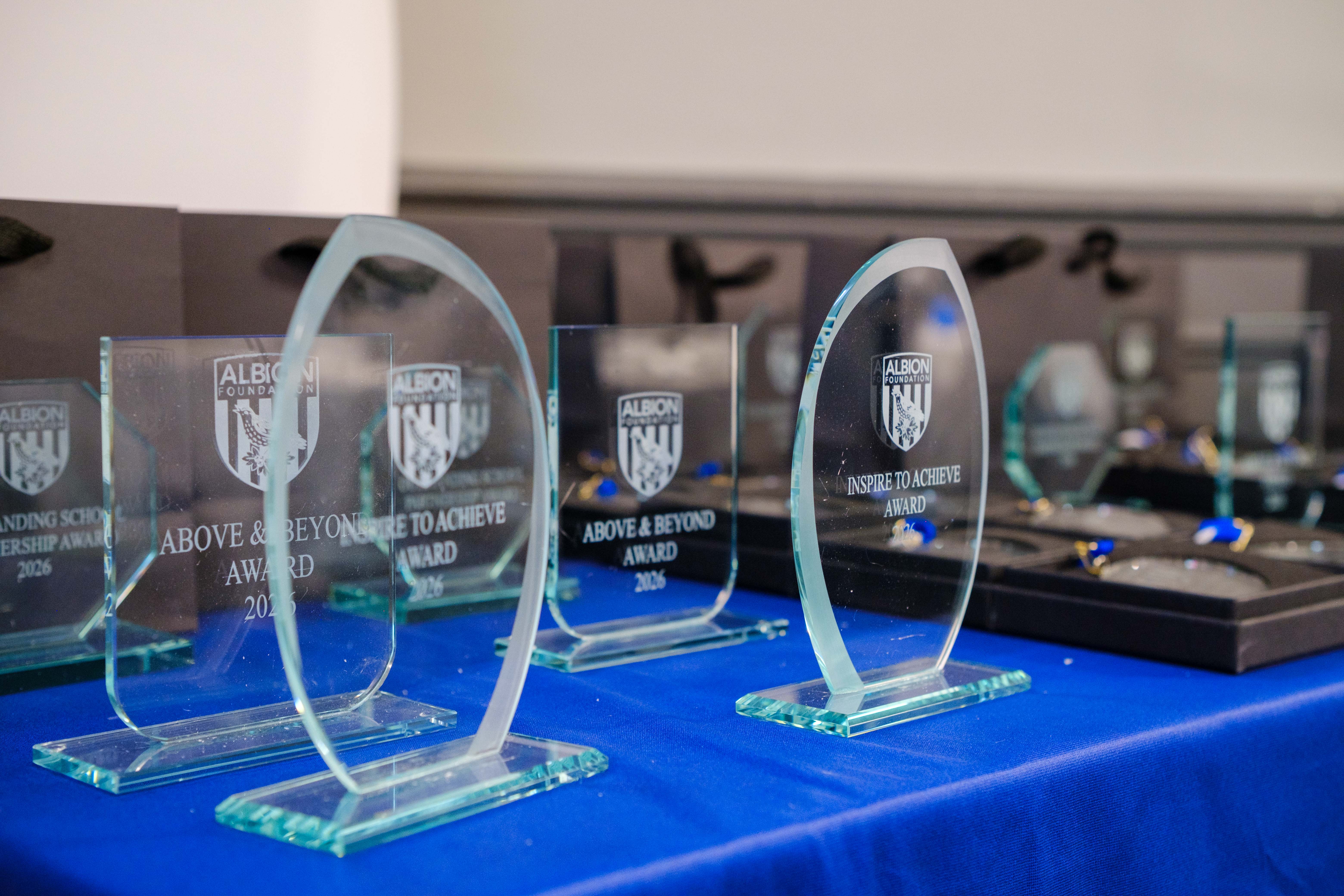Glass trophy awards displayed on a blue-covered table at an Albion Foundation awards event. The engraved awards include categories such as “Above & Beyond Award” and “Inspire to Achieve Award,” with medals and additional trophies visible in the background.
