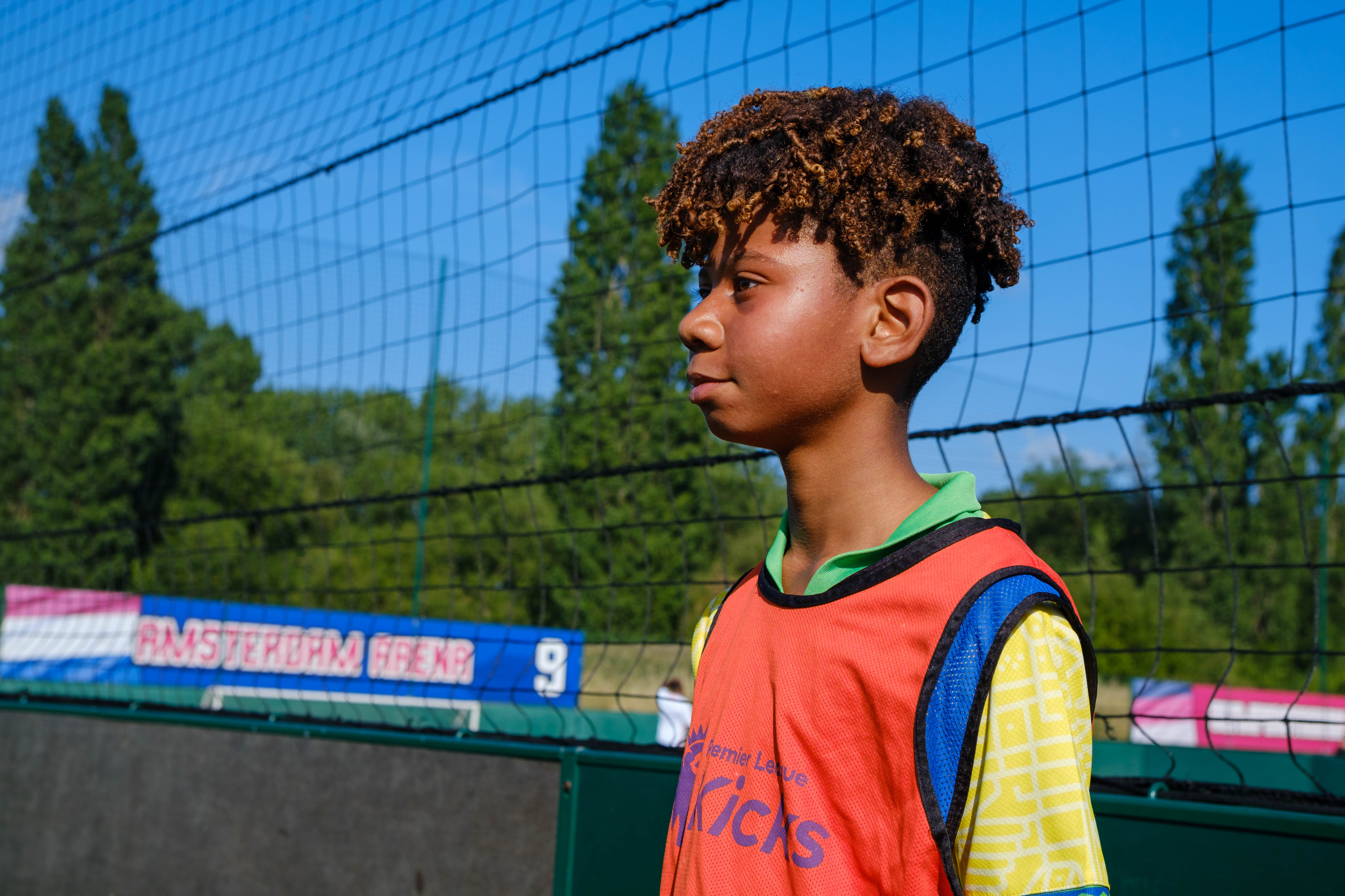 Premier League Kicks participant stands beside a small-sided football pitch, wearing a training bib with goal nets and trees visible in the background.