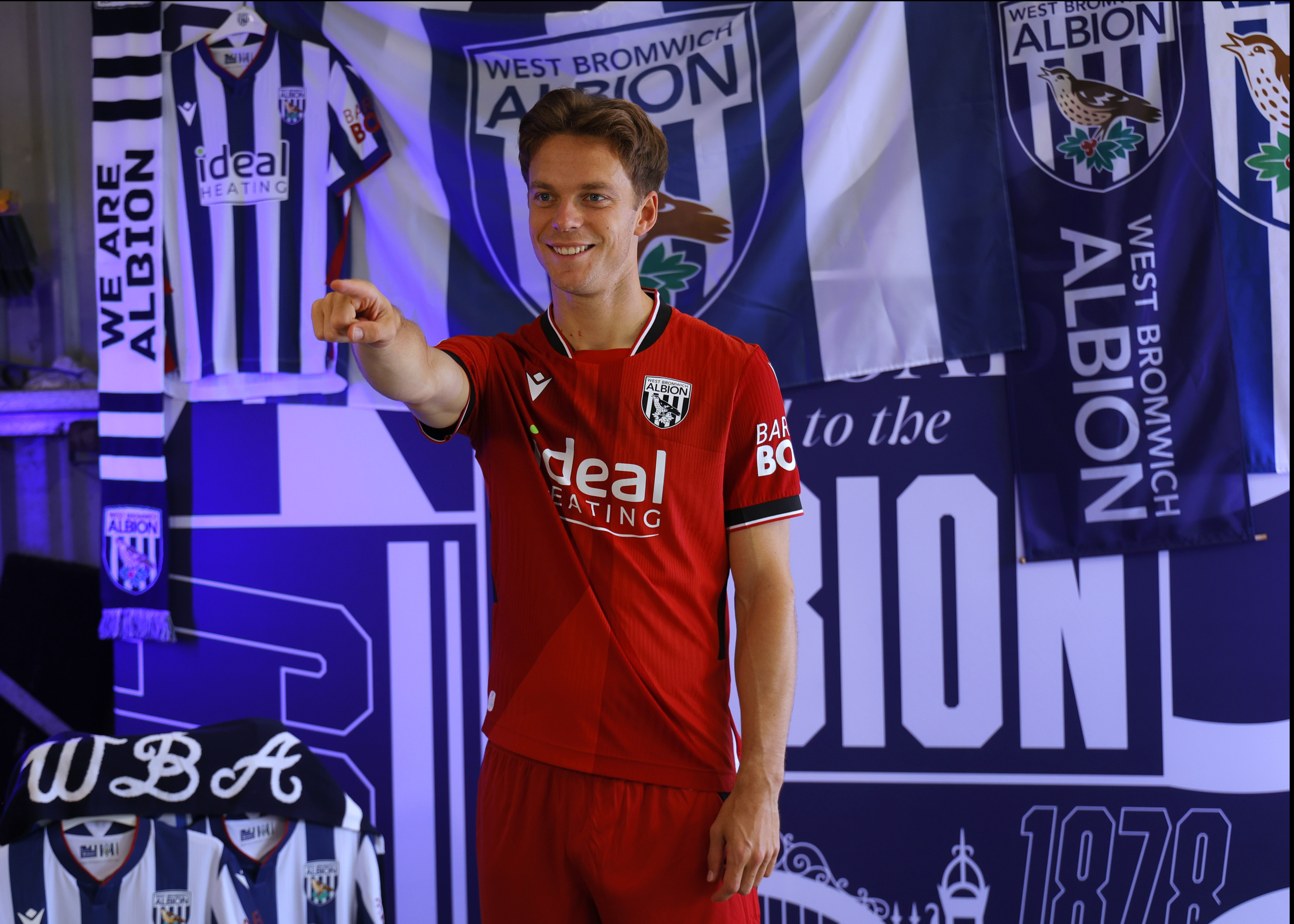 Felix Horn Myhre pointing and smiling at the camera while in the away kit stood in front of WBA branding