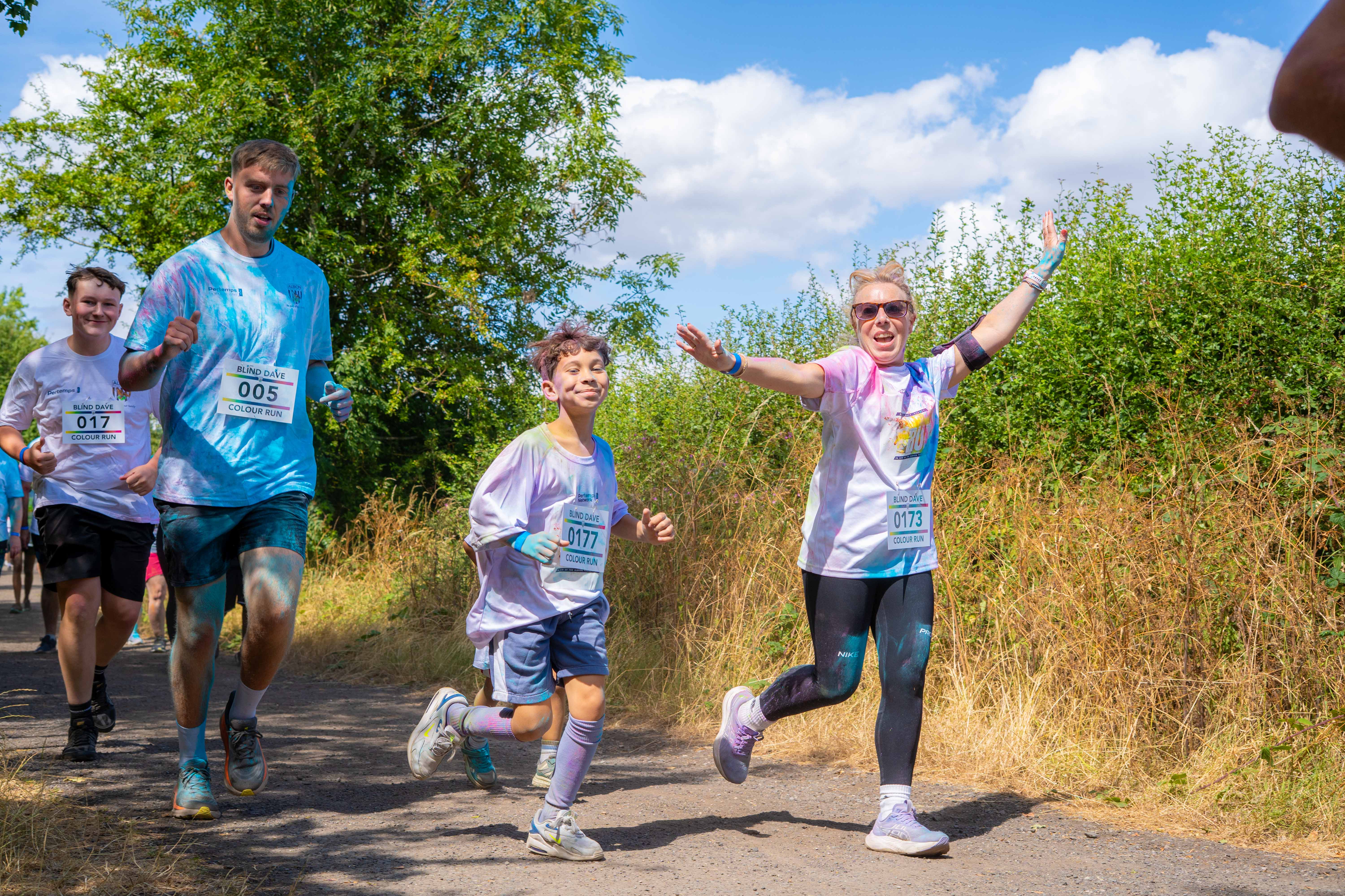Participants running and smiling along a path tall grass and trees.