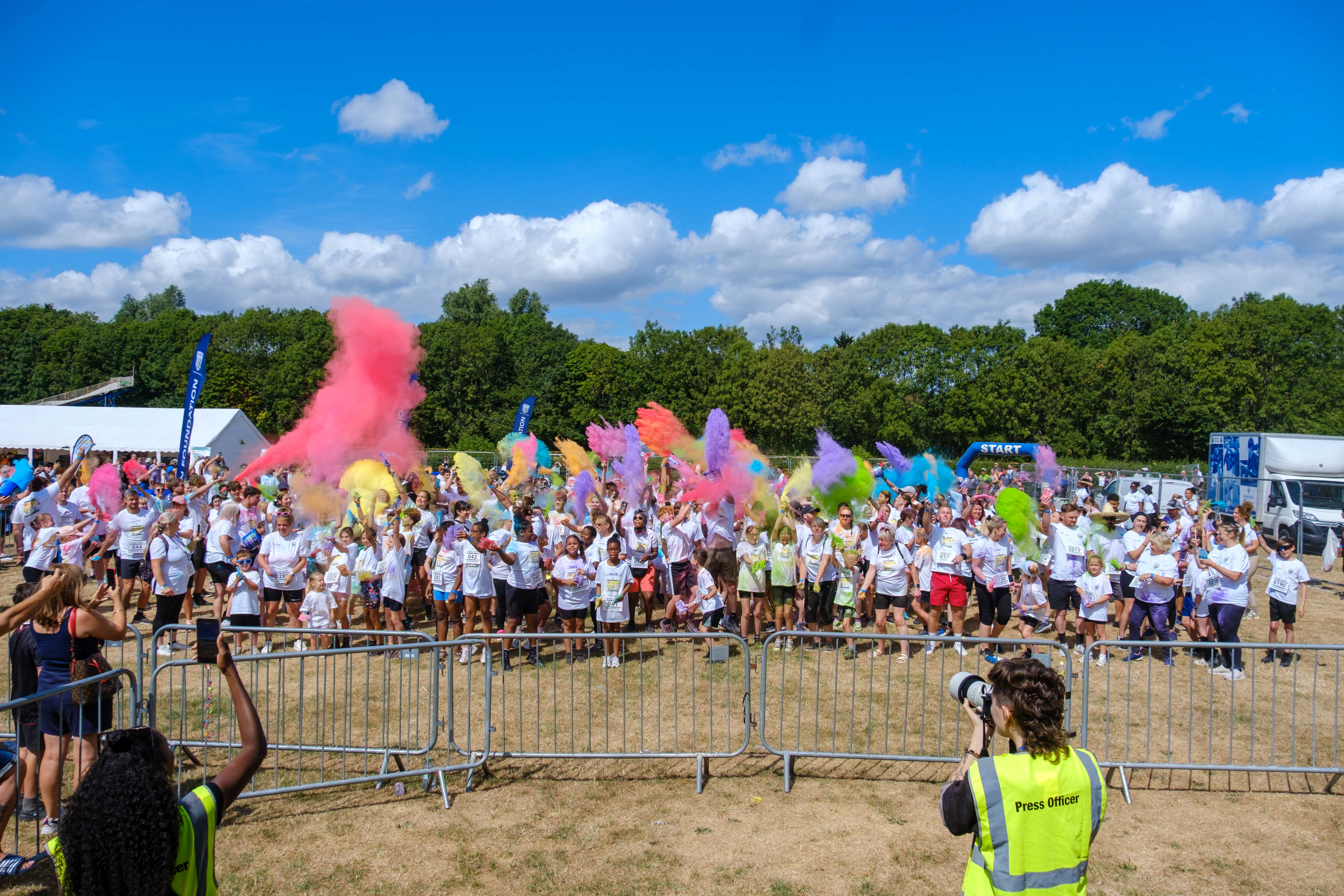 Participants throw brightly coloured powder into the air during a charity colour run, creating large clouds of pink, purple, blue, yellow and green across an open field.