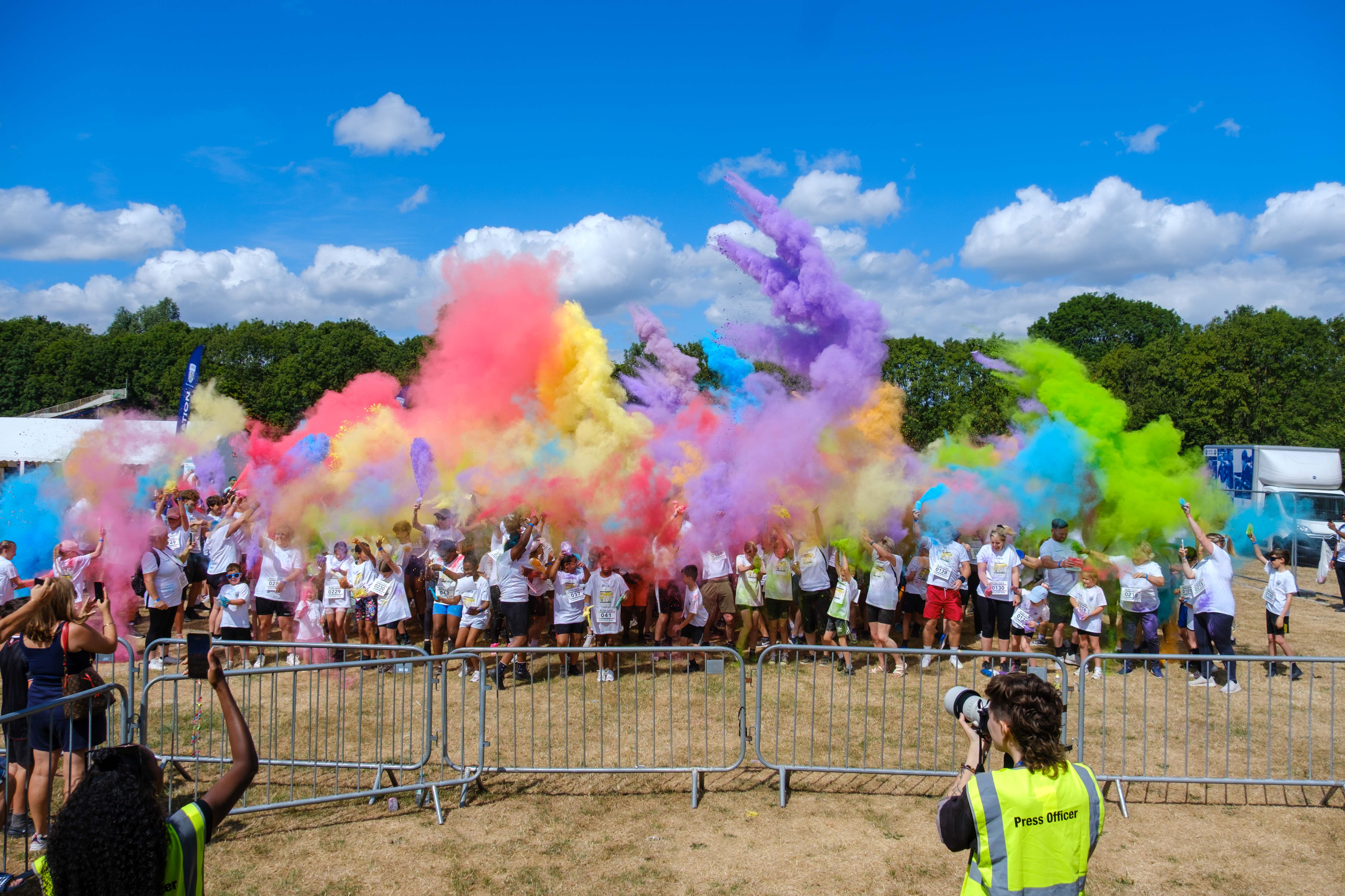 Participants throw brightly coloured powder into the air during a charity colour run, creating large clouds of pink, purple, blue, yellow and green across an open field.