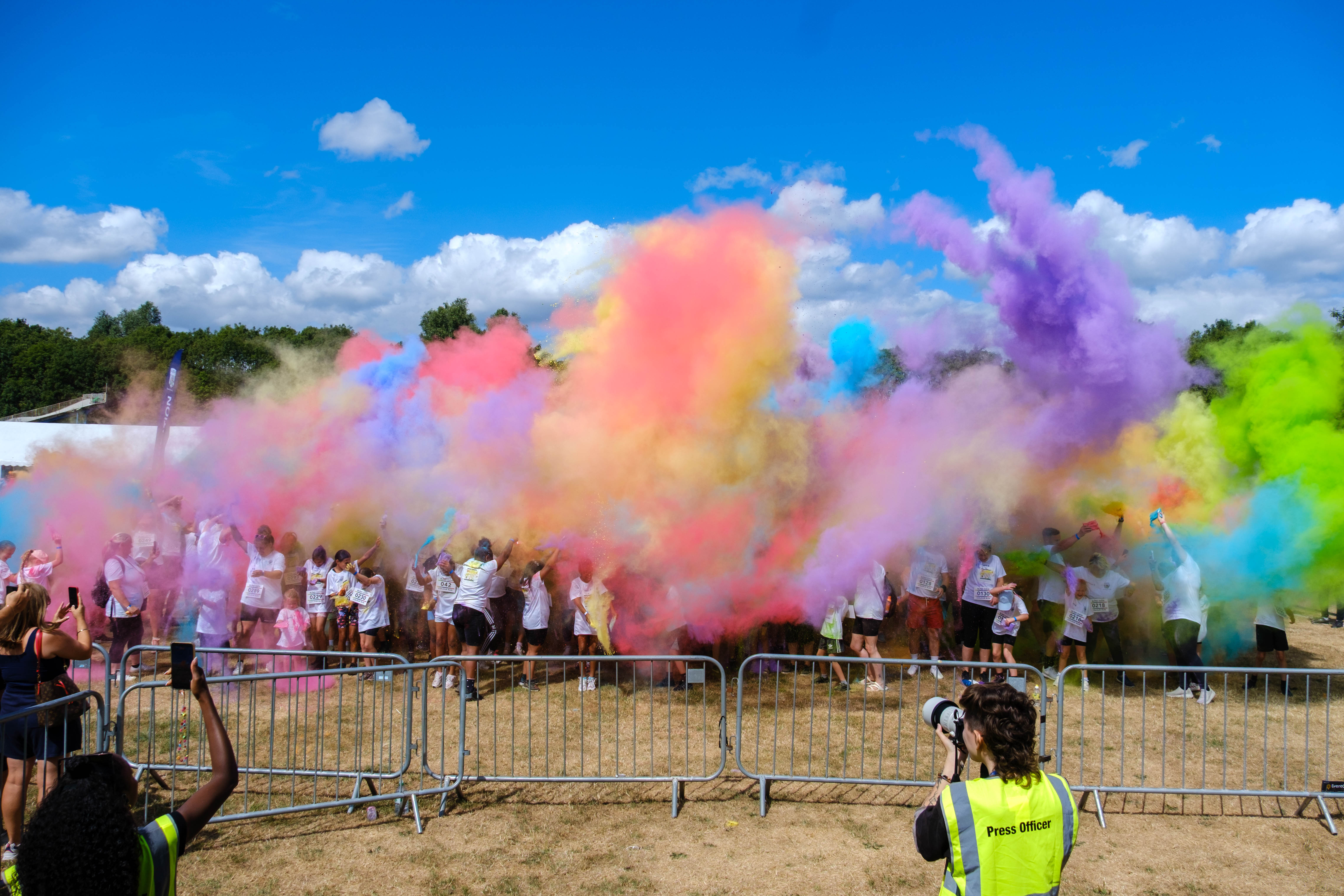 Participants throw brightly coloured powder into the air during a charity colour run, creating large clouds of pink, purple, blue, yellow and green across an open field.