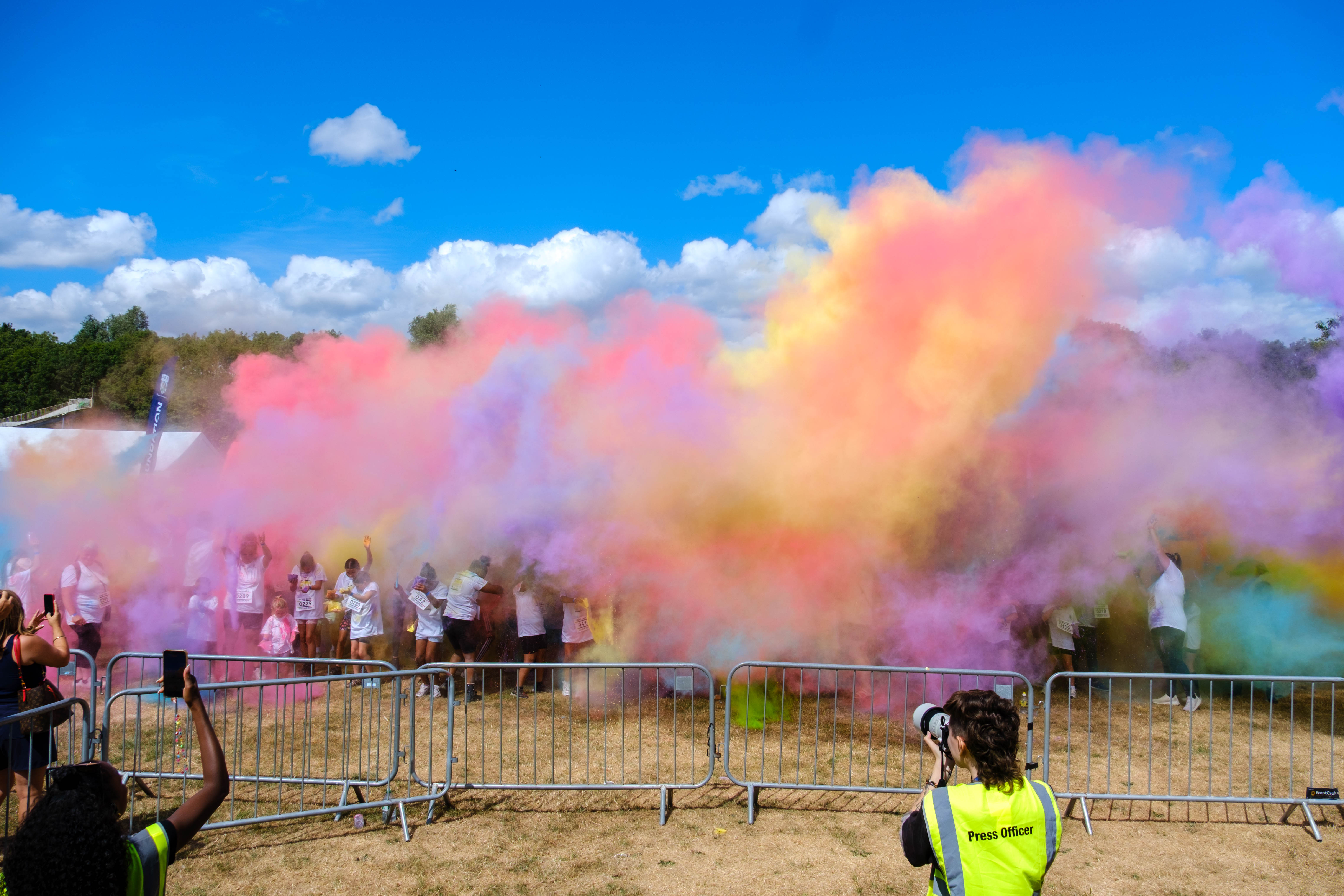 Participants throw brightly coloured powder into the air during a charity colour run, creating large clouds of pink, purple, blue, yellow and green across an open field.