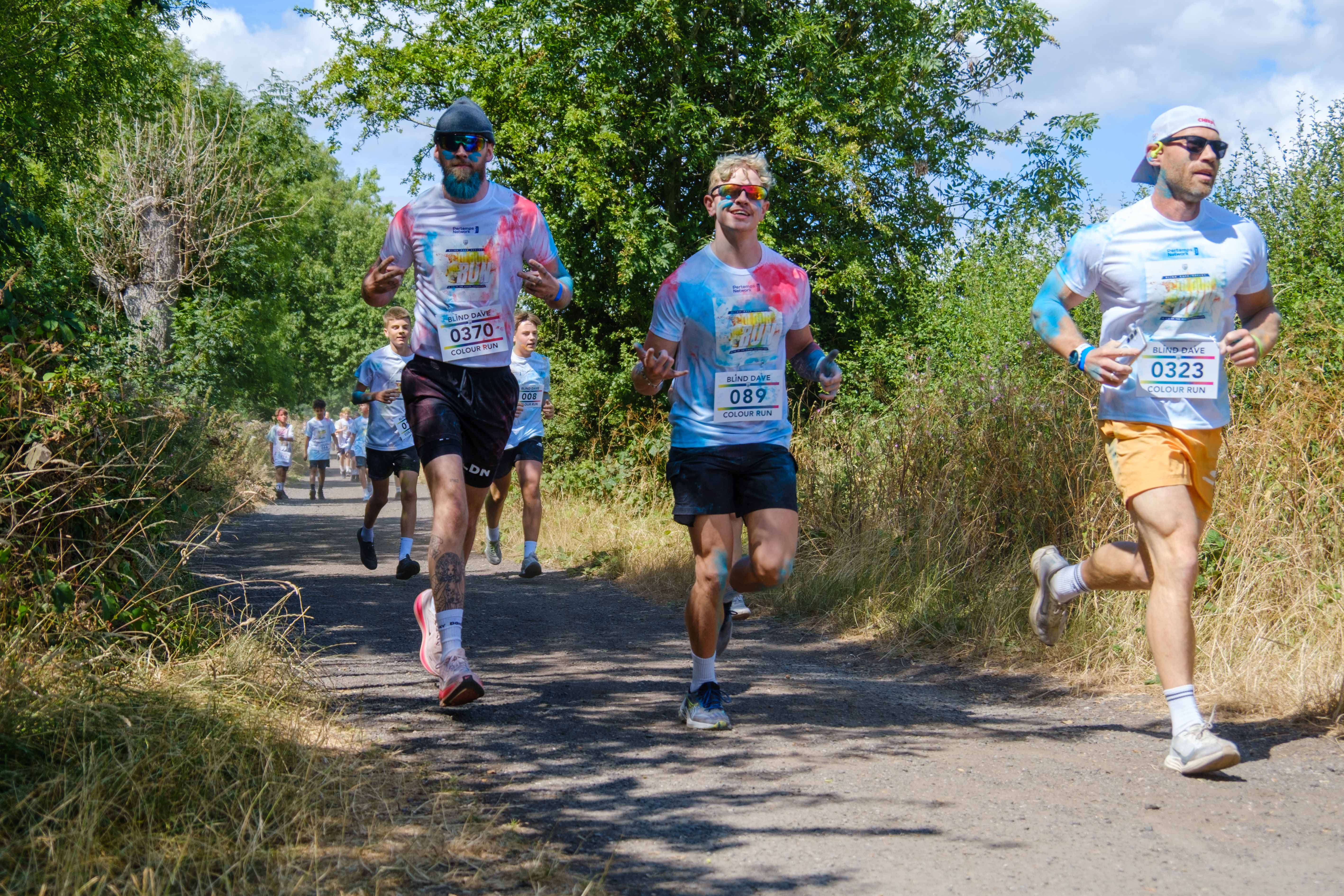 Several participants covered in blue and pink color powder run along a tree-lined countryside path