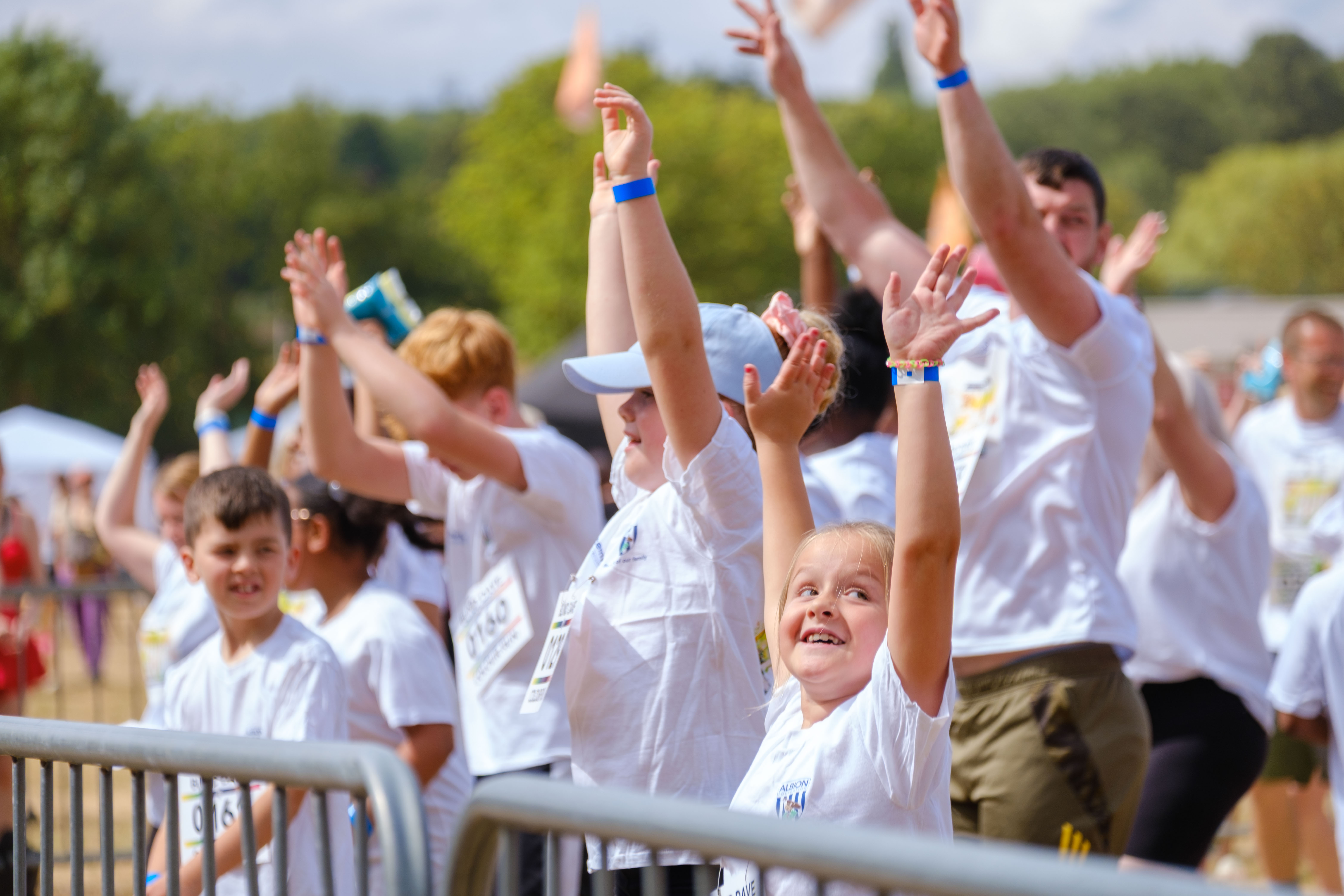 Colour run participants raise their hands during a warm-up session behind barriers, wearing event shirts and wristbands before the start of the run.