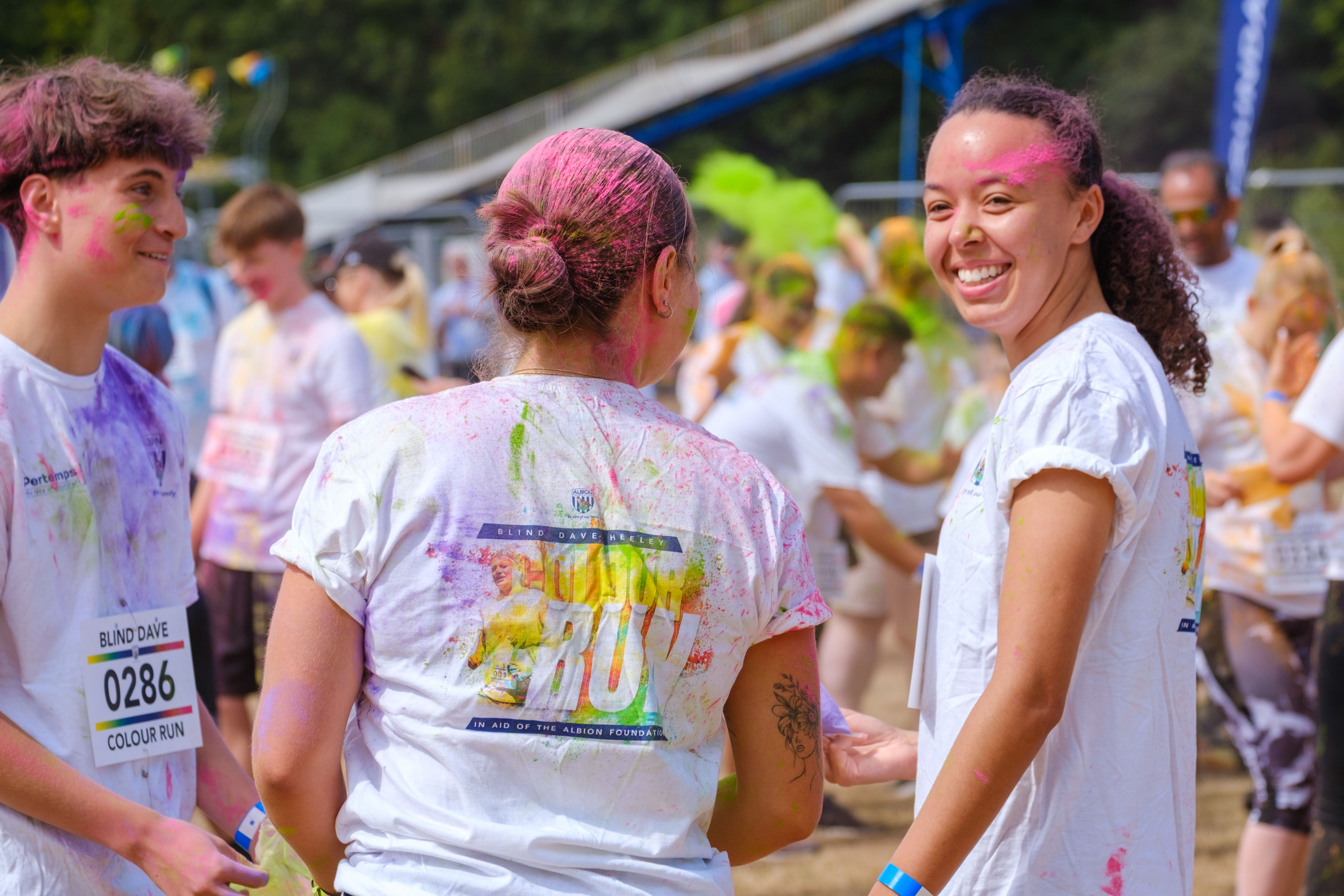 Colour run participants covered in brightly coloured powder gather on an open field, wearing event shirts and bibs during the Blind Dave Colour Run.
