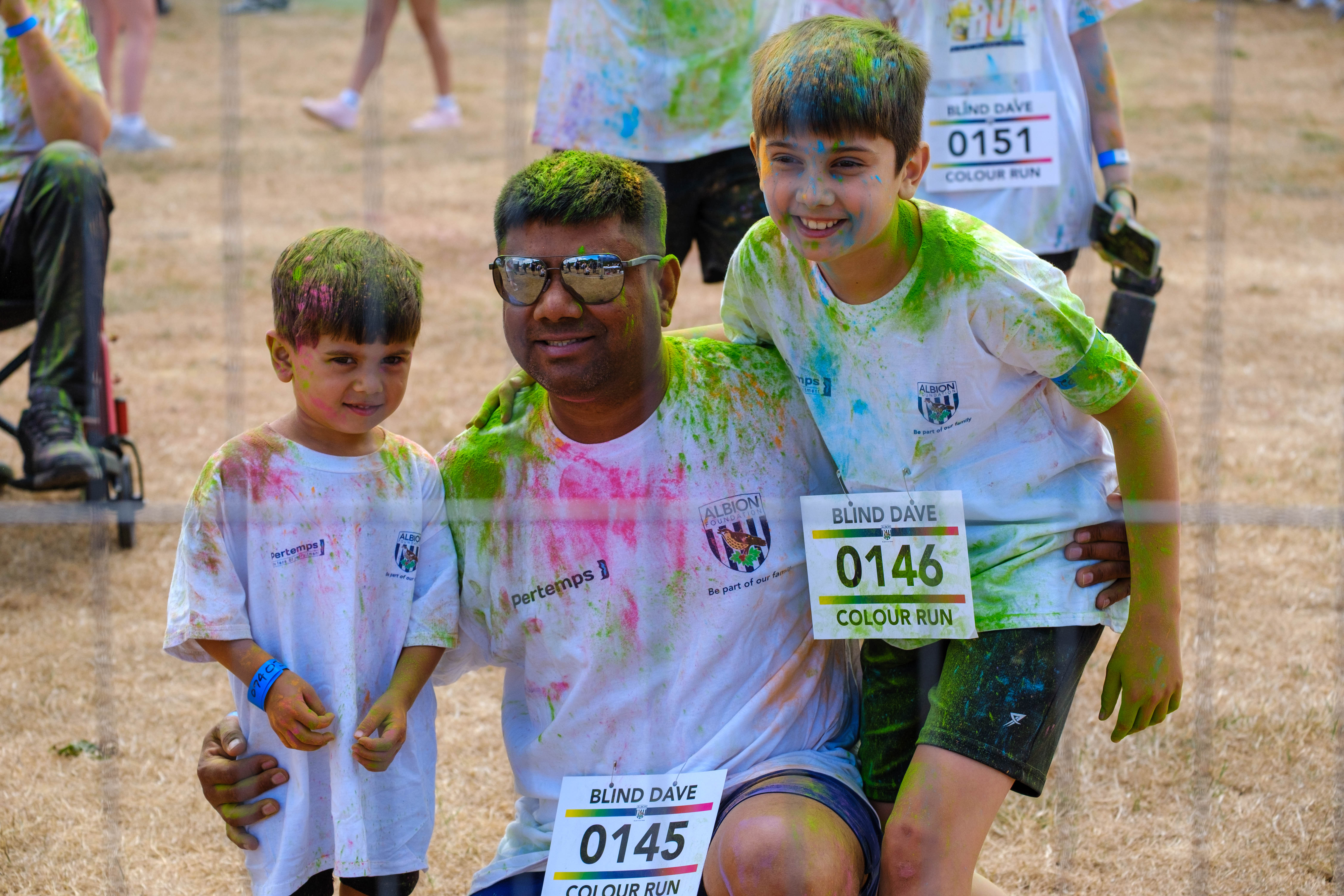 Adult and two children pose together after the Blind Dave Colour Run, covered in coloured powder and wearing event shirts and numbered bibs.