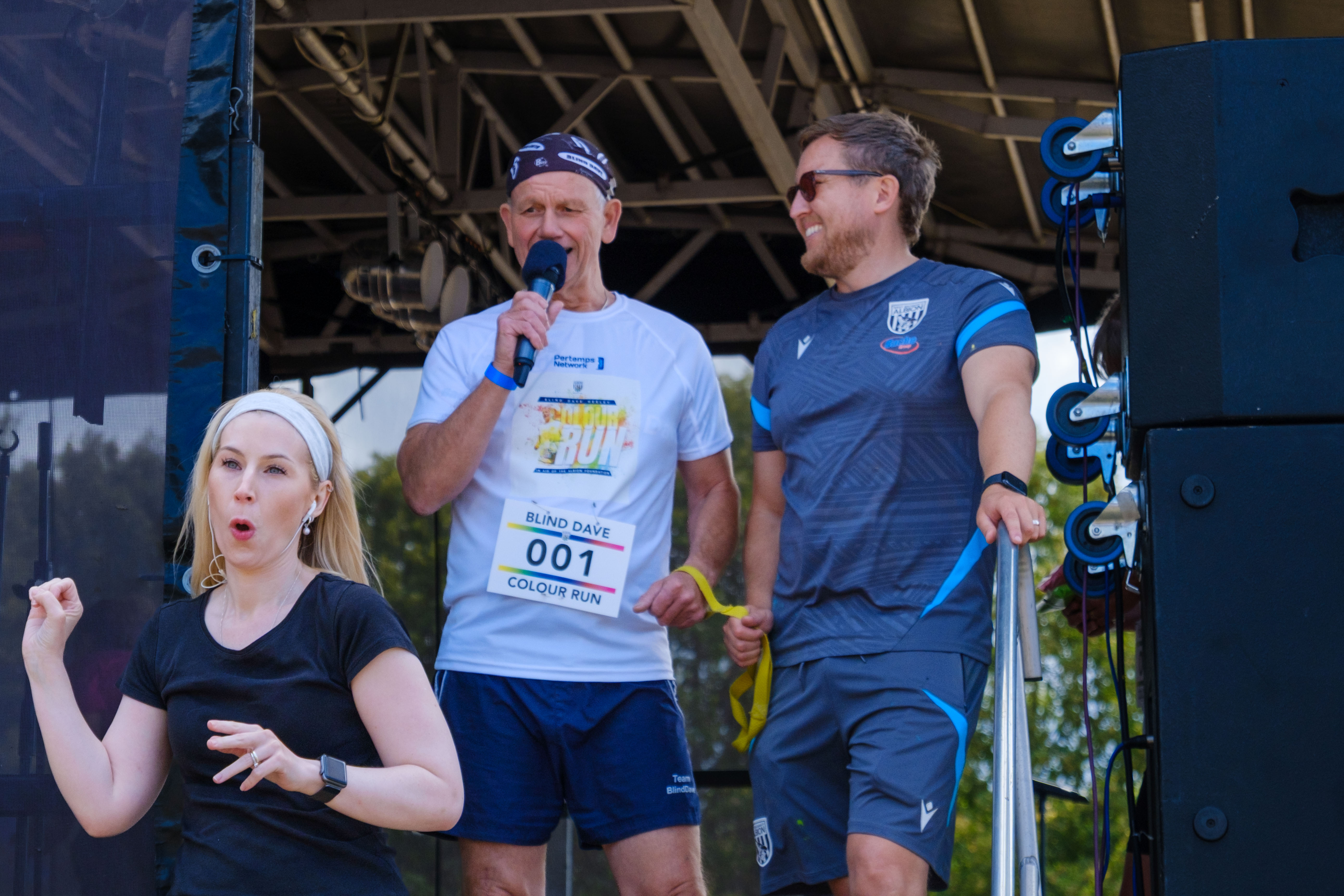 Blind Dave speaks on stage with a microphone before the charity colour run, alongside an Albion Foundation representative, while a British Sign Language interpreter stands in front of the stage.