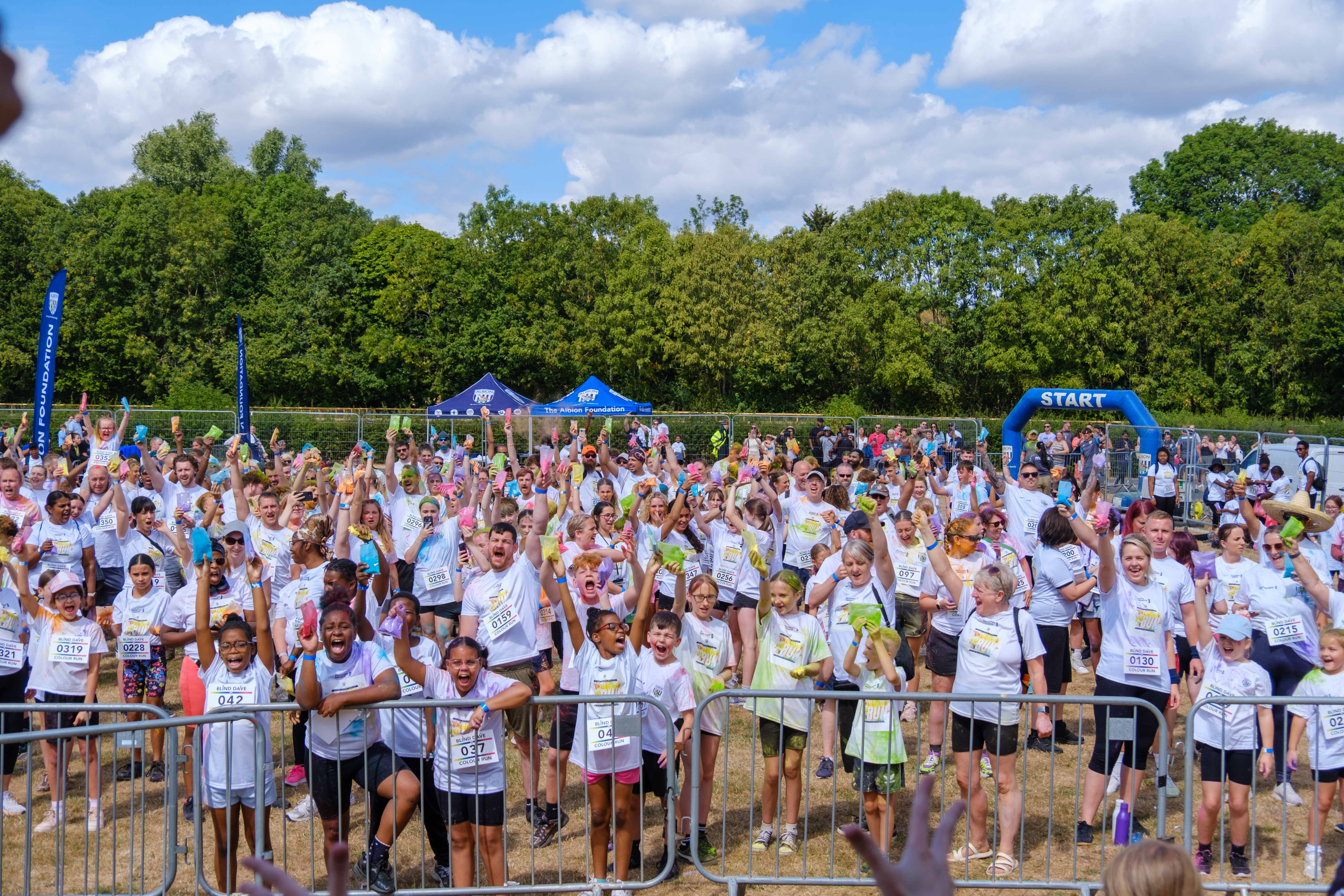 Large group of charity colour run participants gather behind barriers before the start, raising packets of coloured powder in the air on a sunny field, with event tents and a START arch visible in the background.