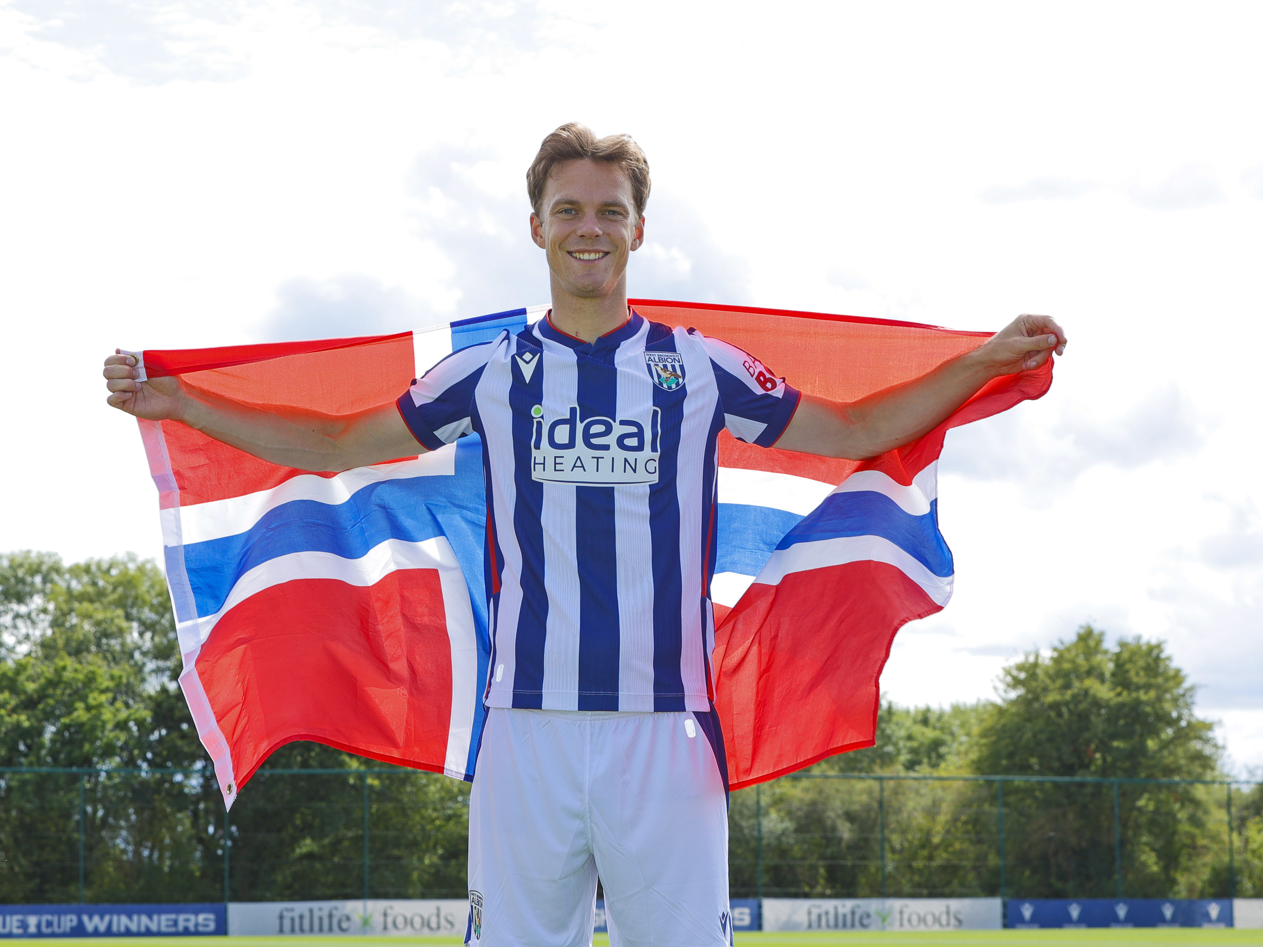 Felix Horn Myhre smiling at the camera while in the home kit with a Norway flag behind him