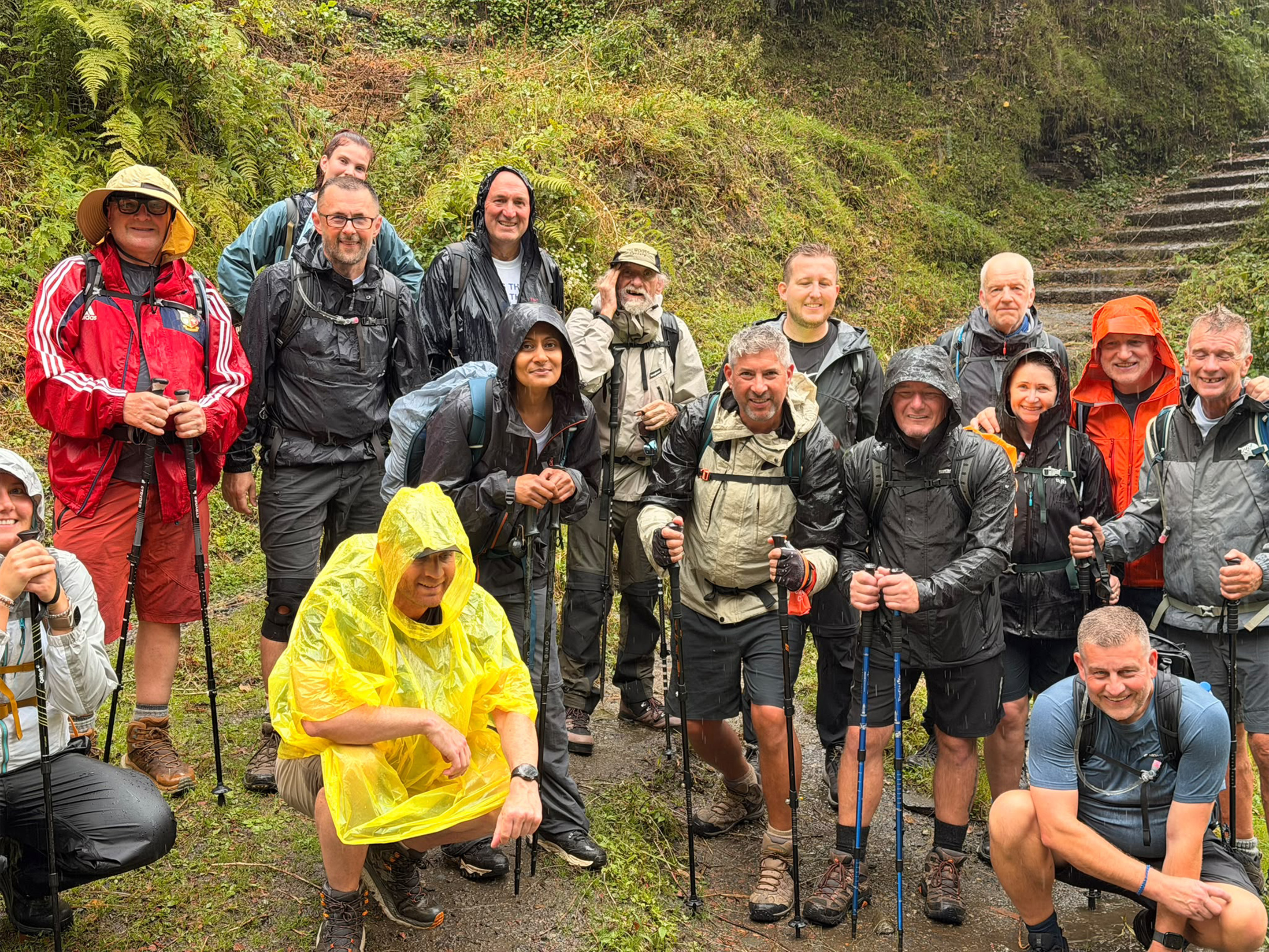 Dalai Lama Himalayan Trek participants in waterproof clothing pause for a group photo on a rain-soaked woodland trail, with stone steps and dense greenery visible behind.