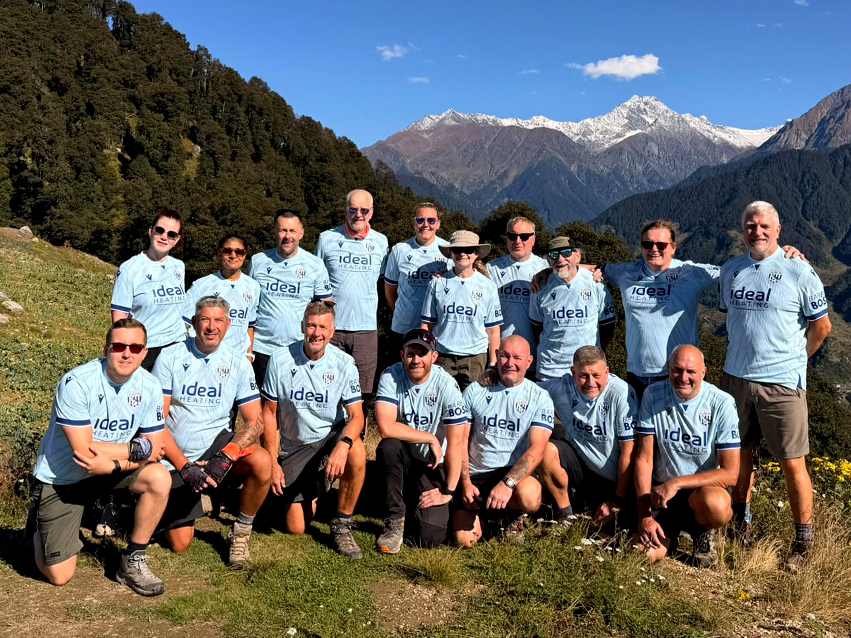 Dalai Lama Himalayan Trek participants wearing matching Albion Foundation trek shirts pose for a group photo on a mountain ridge, with snow-capped Himalayan peaks and forested valleys forming the backdrop under clear blue skies.