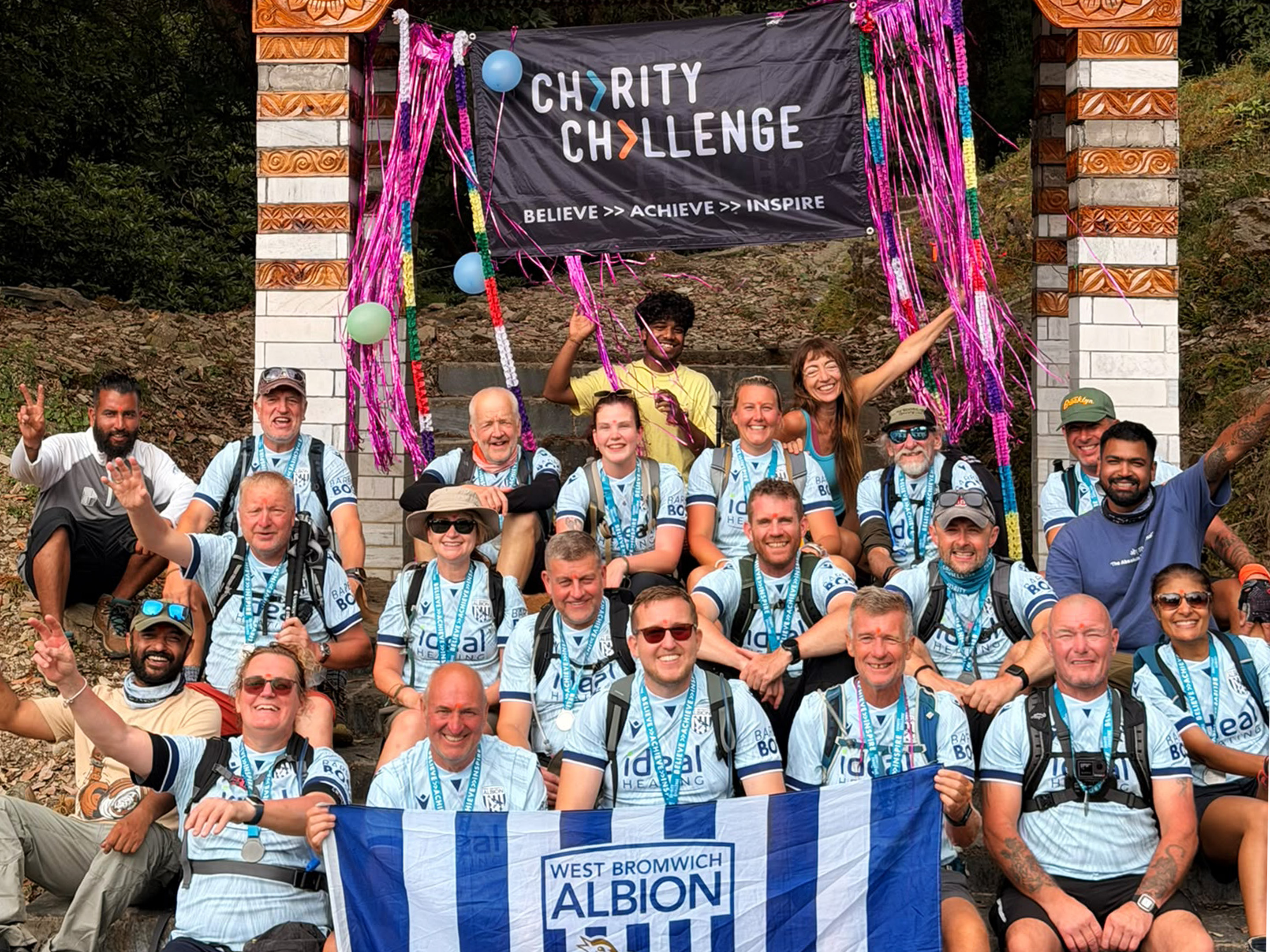 Dalai Lama Himalayan Trek participants gather beneath a “Charity Challenge” finish banner, wearing medals and holding a West Bromwich Albion flag after completing the trek.