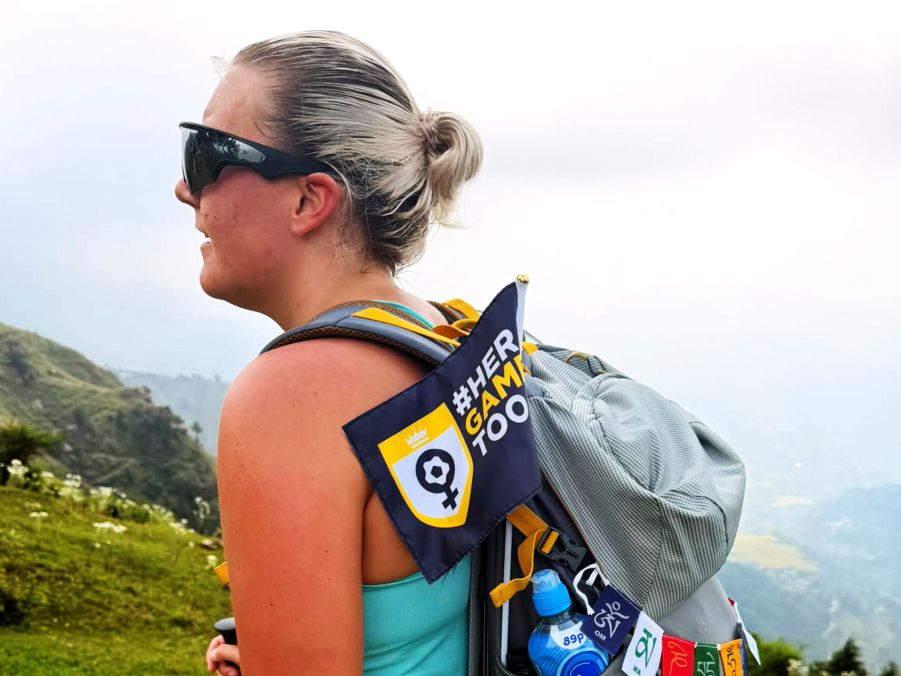 Participant on the Dalai Lama Himalayan Trek stands on a mountainside wearing sunglasses and carrying a backpack decorated with a “#HerGameToo” flag and colourful prayer flags. Rolling hills and misty mountain scenery stretch into the distance behind them.