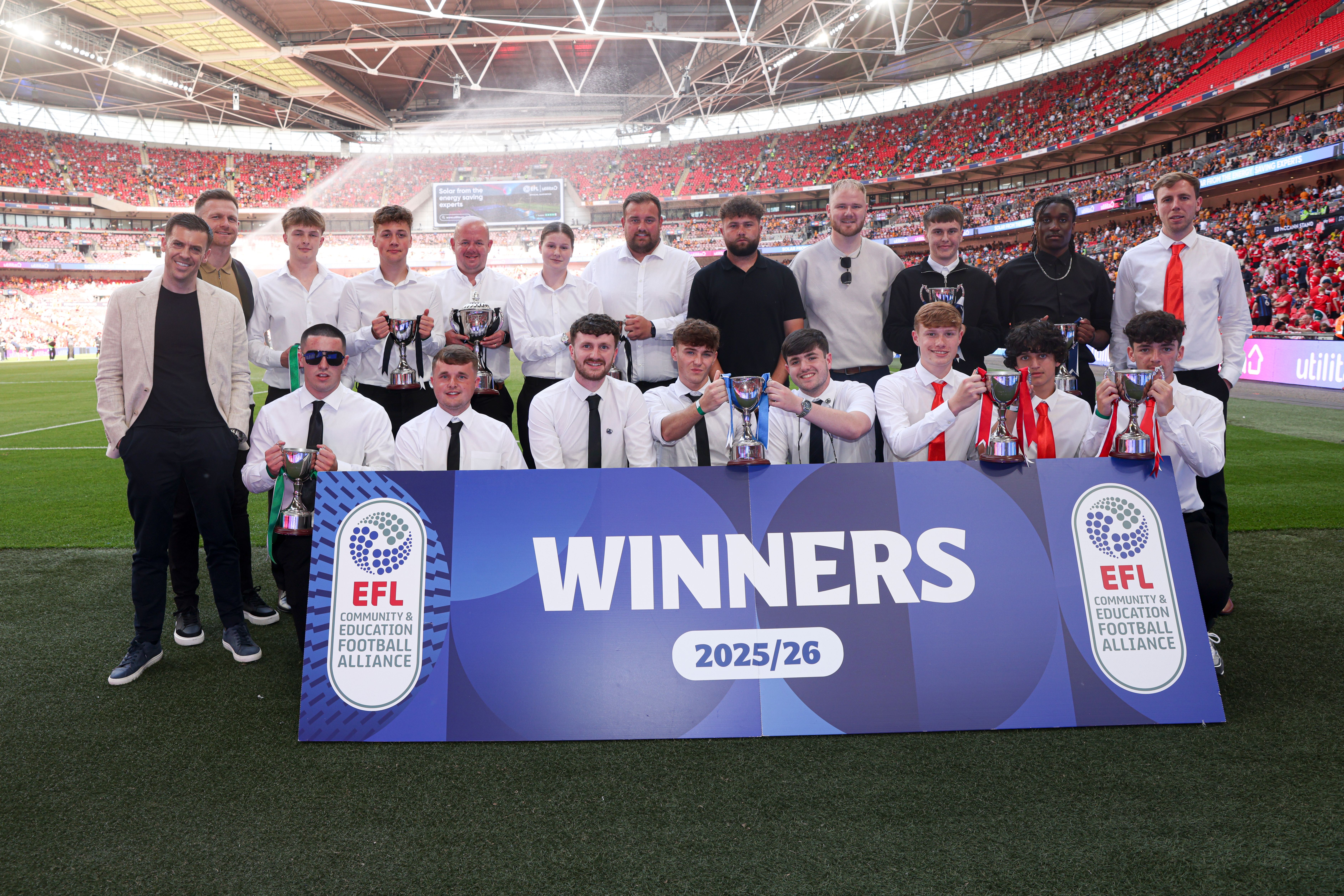 Post-16 team at Wembley behind a Winners banner holding trophies.
