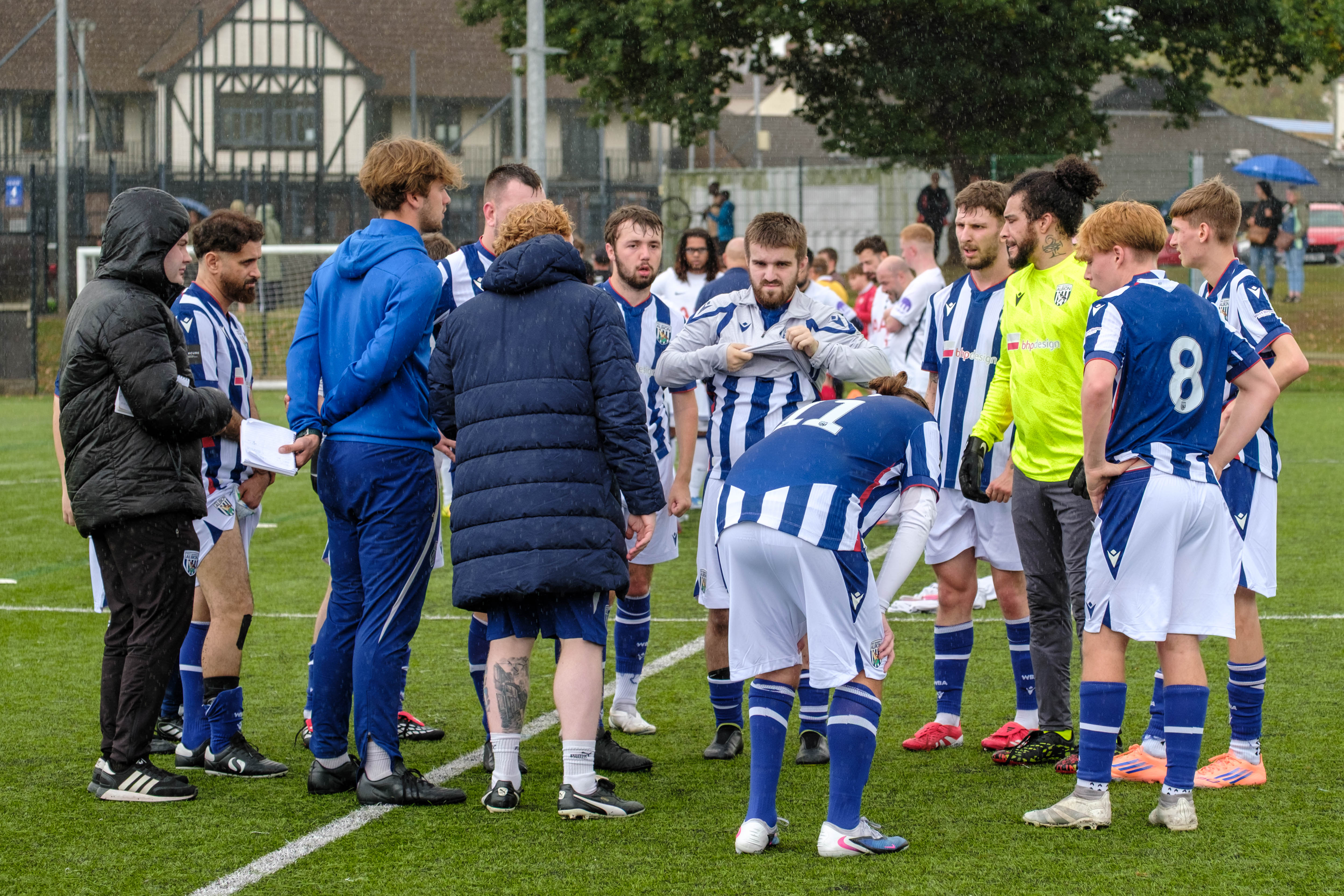 West Bromwich Albion Cerebral Palsy players and coaches gather in a huddle on an outdoor football pitch during a break in play, with light rain falling and opposing players visible in the background.