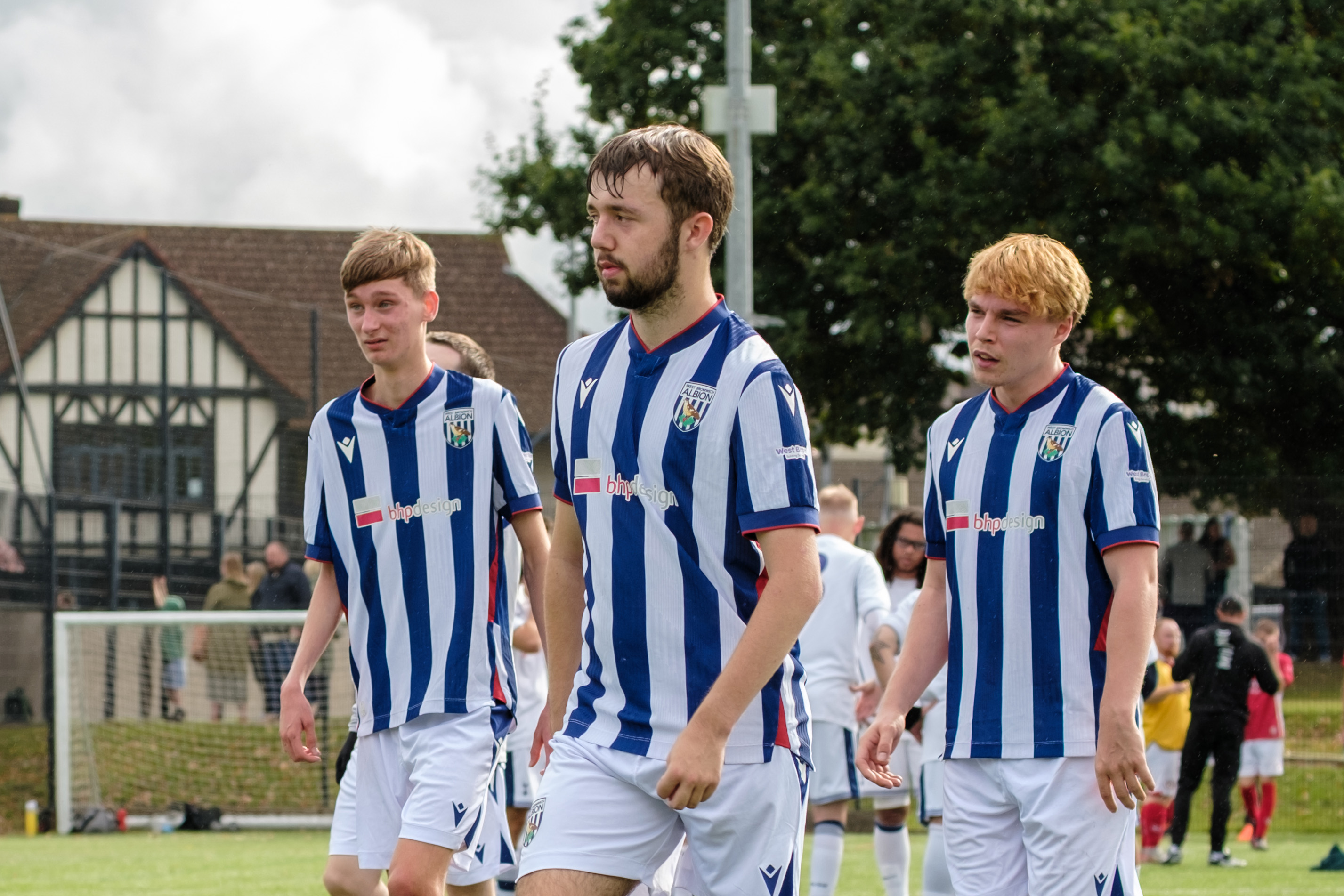 Three West Bromwich Albion Cerebral Palsy players in blue-and-white striped kits walk across an outdoor football pitch during a match, with teammates, spectators and a goal visible in the background.