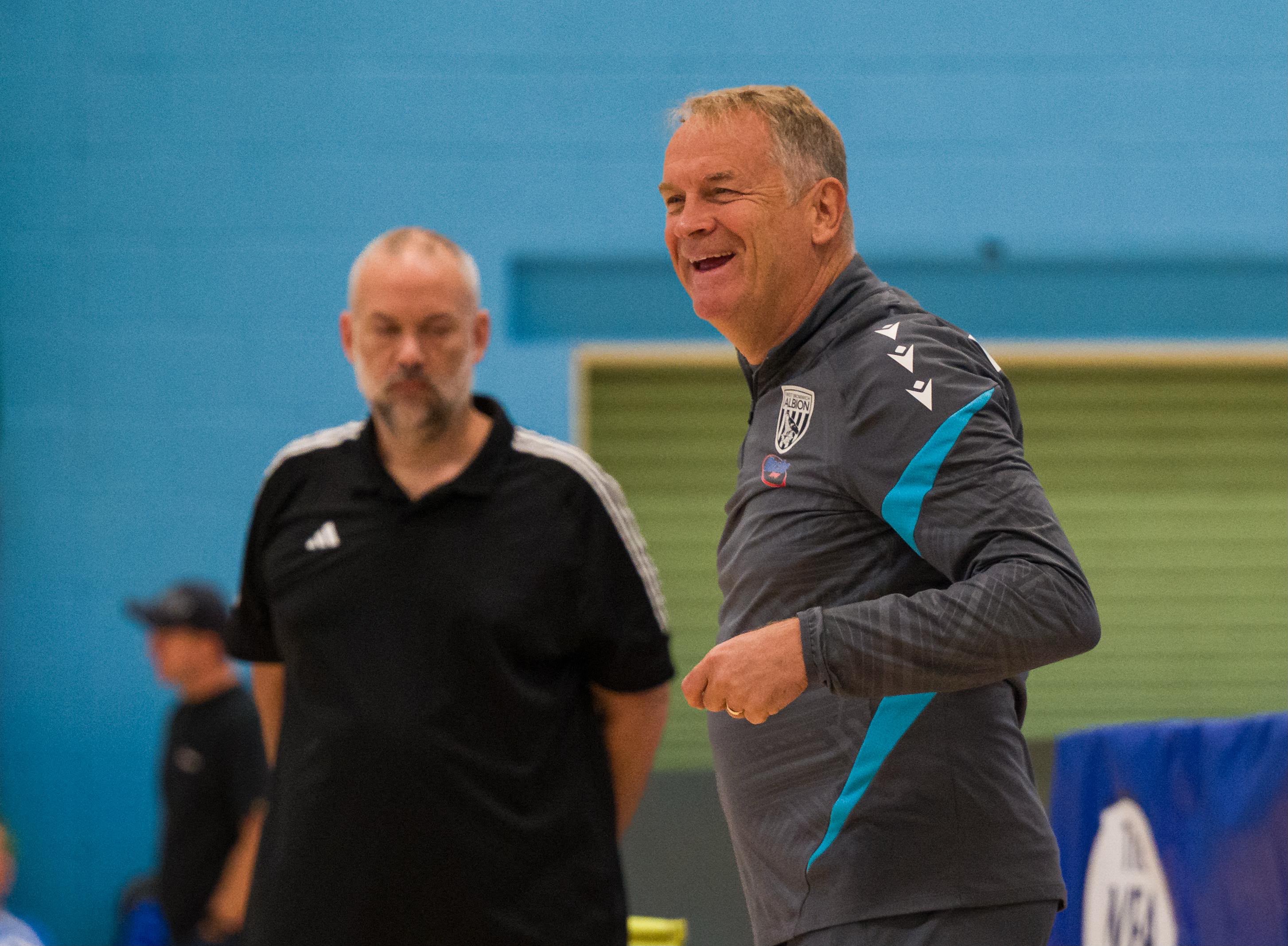 Coach Colin Gordon standing courtside during an indoor powerchair football match, wearing West Bromwich Albion training kit, with another official and sports hall equipment visible in the background.